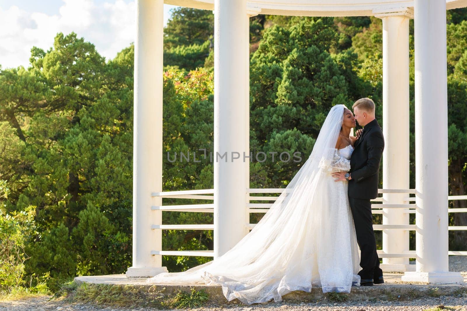 Portrait of mixed-racial newlyweds against the background of a gazebo with round columns, a girl in a lush white dress