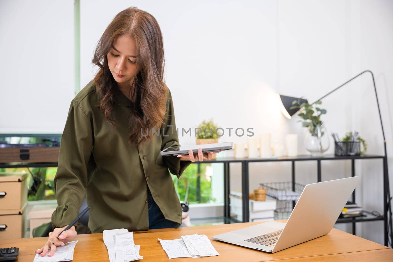Asian female calculating family budget financial and accounting at home, young woman working with financial data calculator with tablet computer and writing make note calculate, pay bills tax online