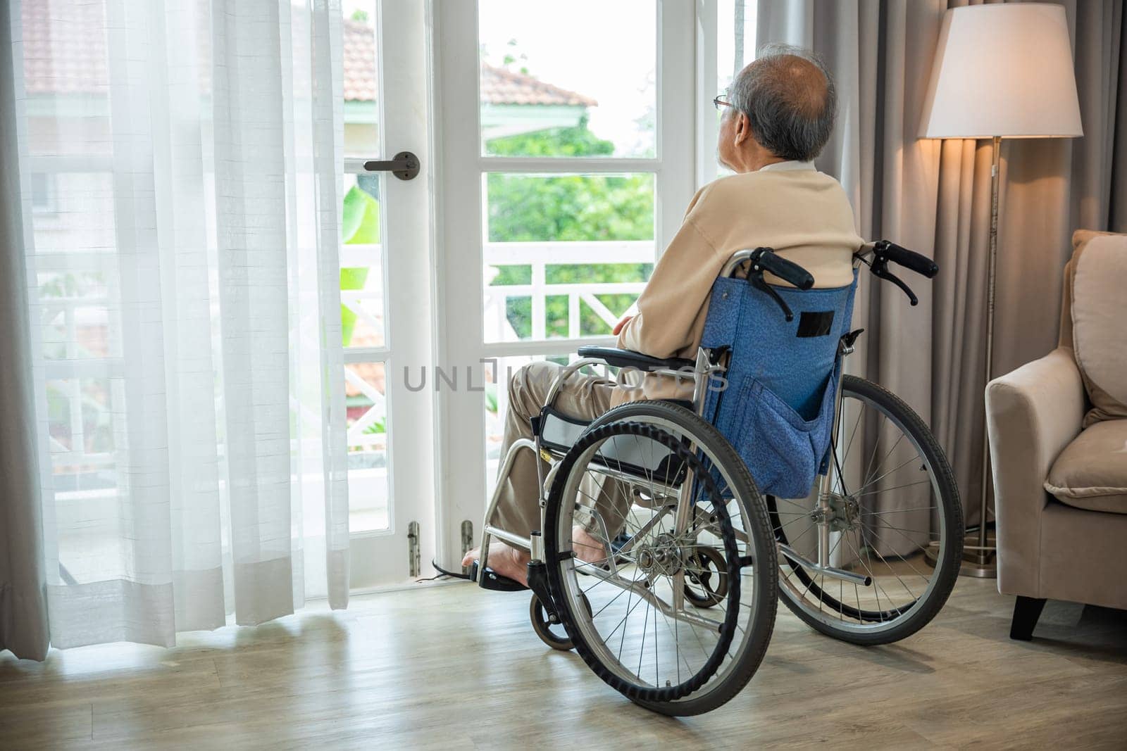 Asian senior man disabled sitting alone in wheelchair looking through window at hospital, lonely elder thoughtful sad old man look outside windows in bedroom at retirement home, feel depressed lonely