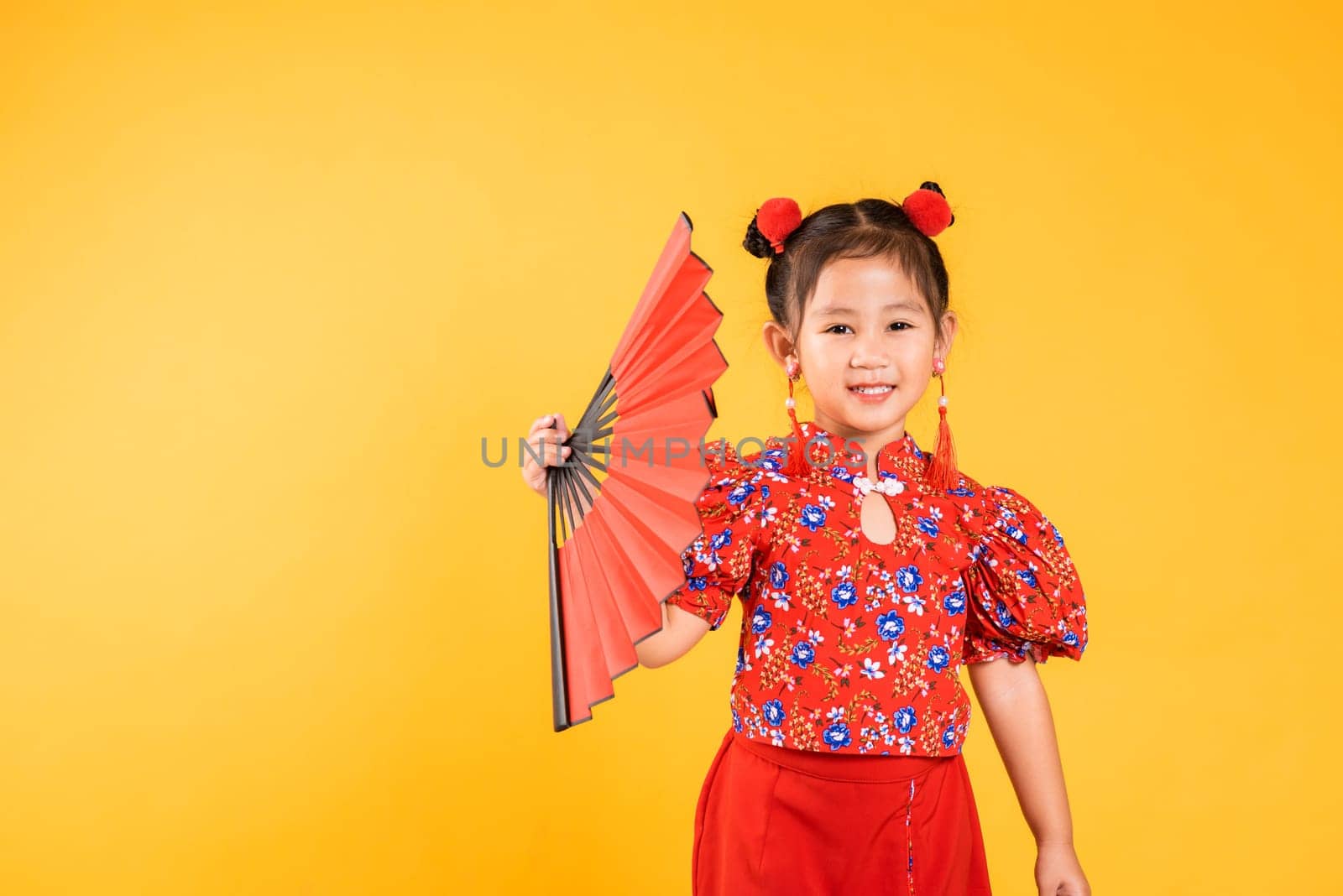 Chinese New Year. Happy Asian Chinese little girl smile wearing red cheongsam holding fan, Portrait children in traditional dress, studio short isolated on yellow background