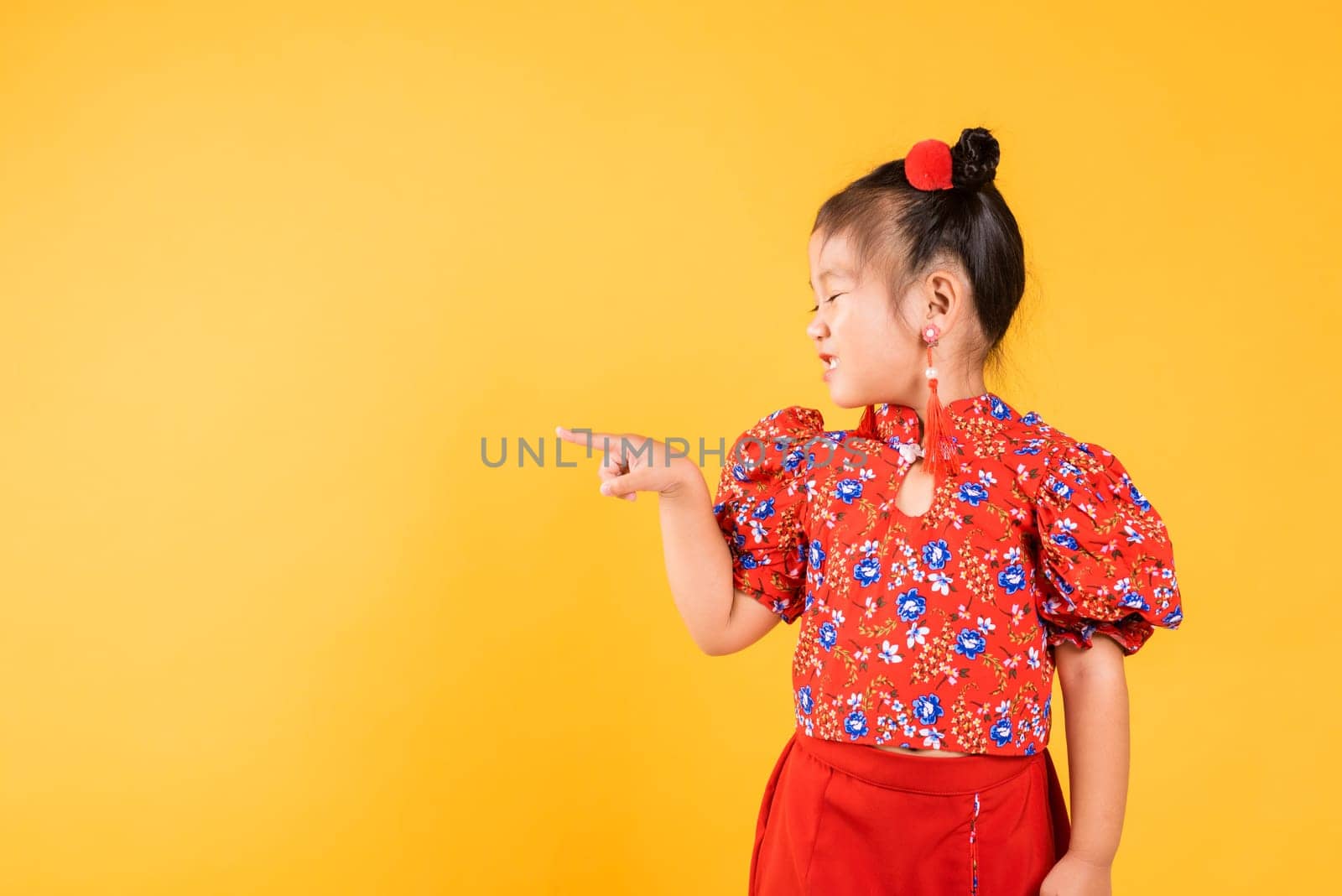 Happy Asian Chinese little girl wear red cheongsam qipao pointing finger to space, Portrait children in traditional dress point to empty, Chinese New Year, studio short isolated on yellow background