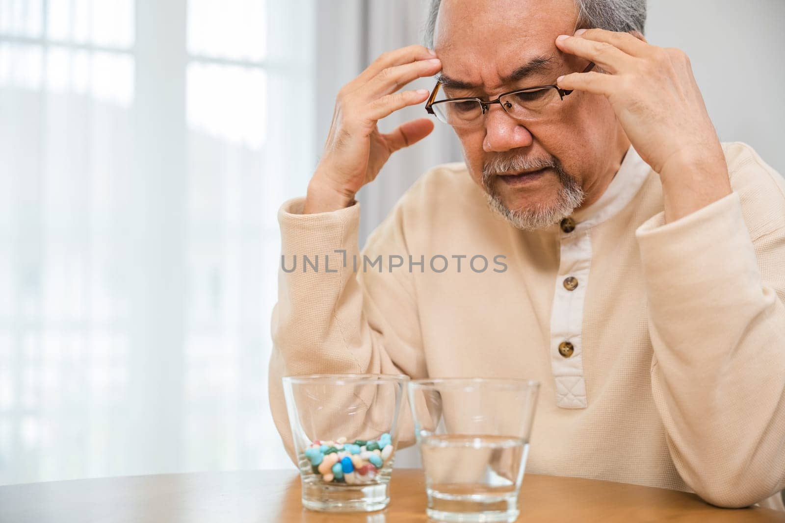 Asian elderly man sick have headache looking pill glass on at home, senior old man grey haired sad sitting in chair hand hold head with many medicines and pills for taking, Elderly healthcare