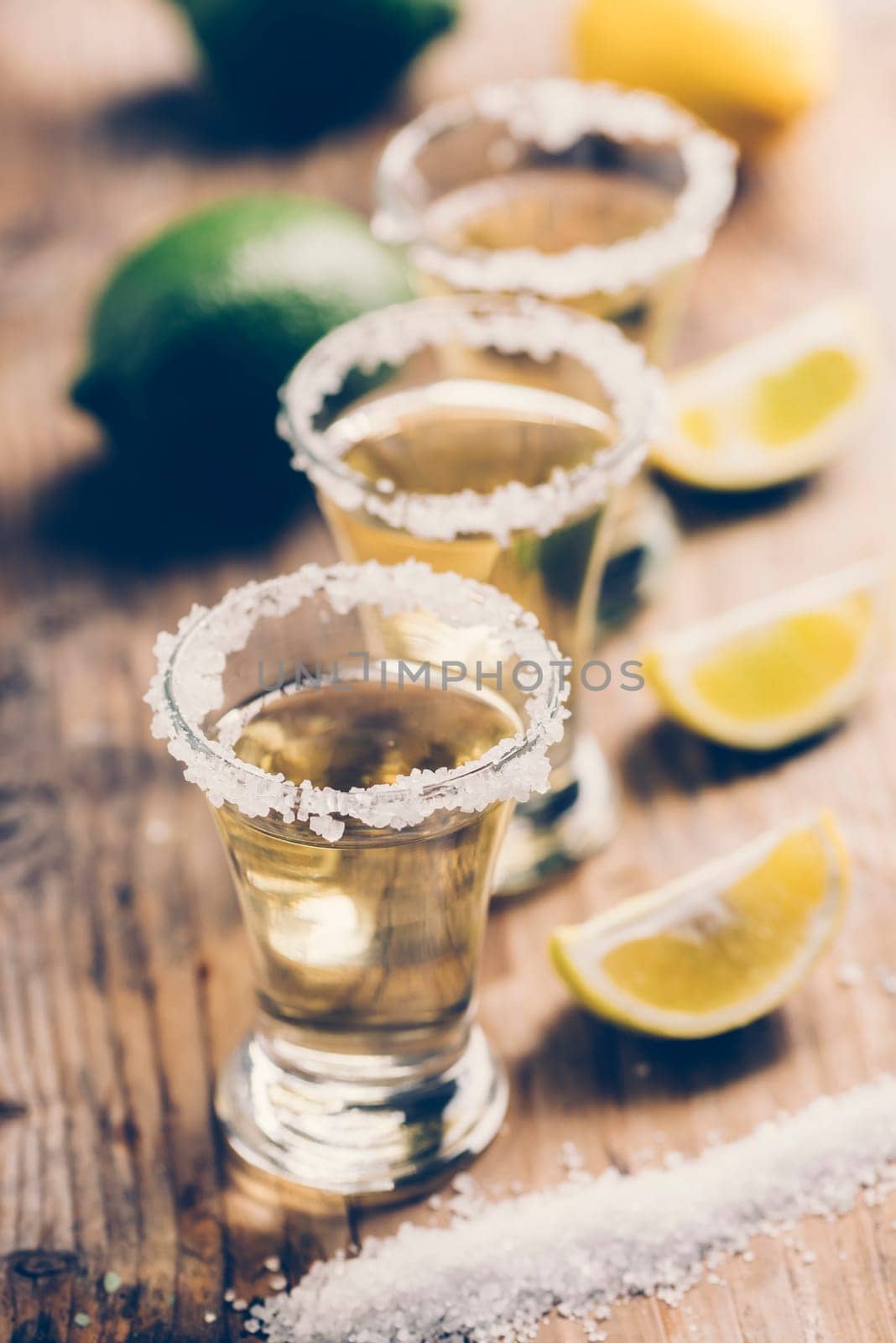 Tree golden mexican tequila shot cocktail with lime slices and salt in row on bar counter with ingredients. Hard liquor glass and lemon on rustic wooden restaurant table. Retro style filter.