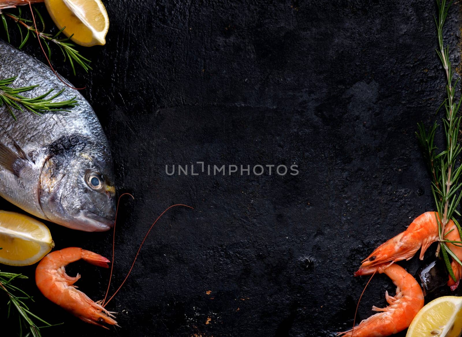 Top view of fresh sea food on grunge textured black background. Shrimps, bream, herbs, lemon and ice on slate with copyspace.