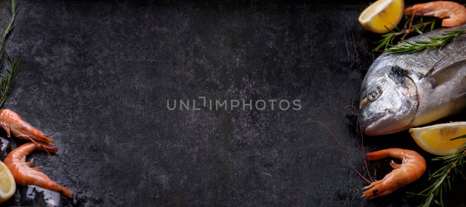 Top view of fresh sea food ingredients on grunge textured black background design. Shrimps, bream sea fish, herbs, lemon and ice on rustic dark slate with copyspace.