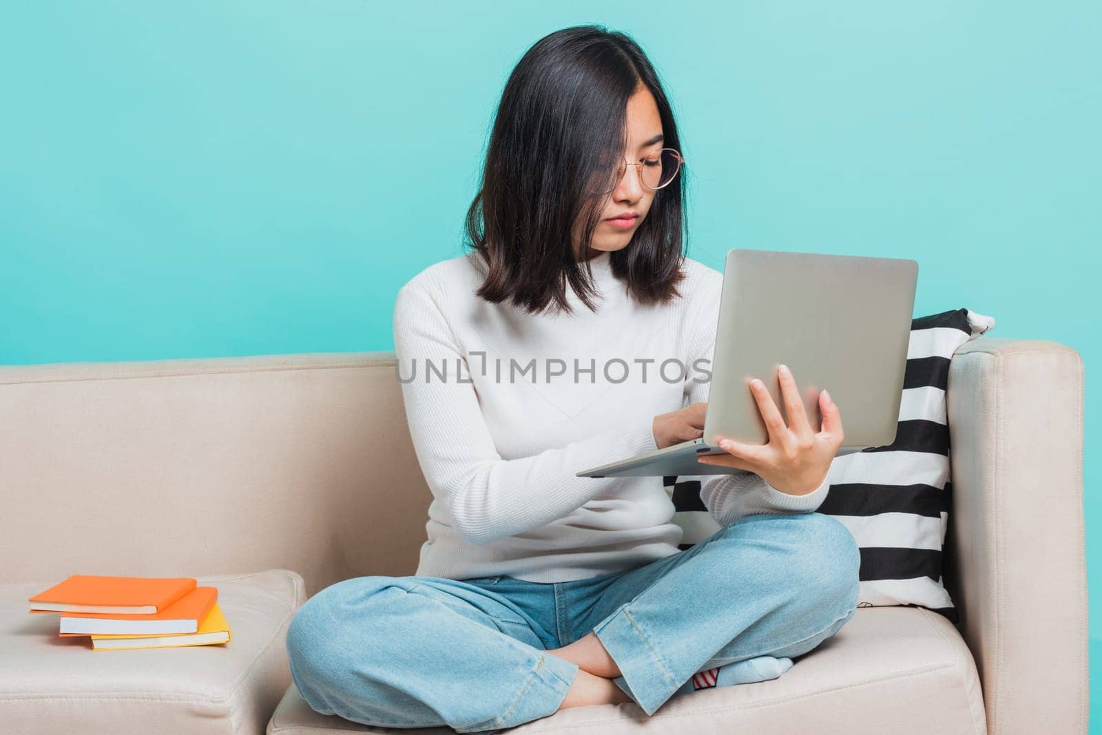 Asian beautiful young woman wear eyeglasses sitting on sofa using a laptop, portrait relaxation of happy female smiling in living freelancer working on computer studio shot isolated on blue background