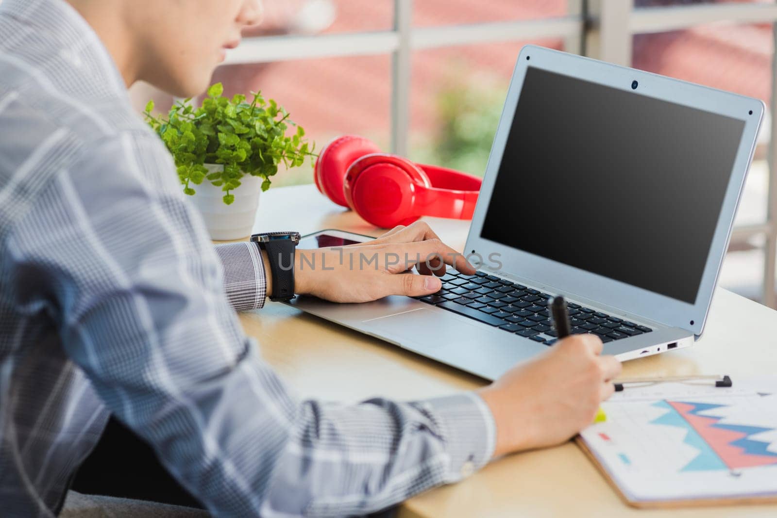 Asian businessman writing notes with pen inspired ideas list on paper notepad while working on laptop computer. Happy handsome man sitting busy on desk table working from home office doing note