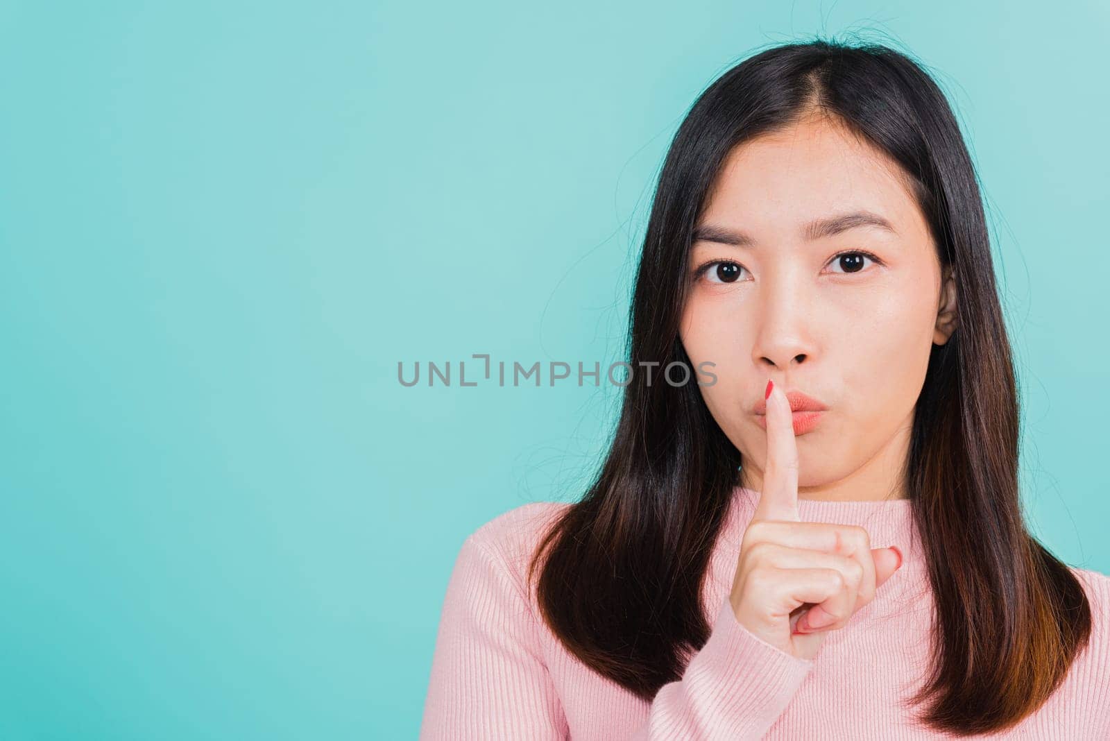 Young Asian beautiful woman with finger in front of mouth on lips, sign of silence gesture saying shh or keeping a secret, studio shot isolated on blue background