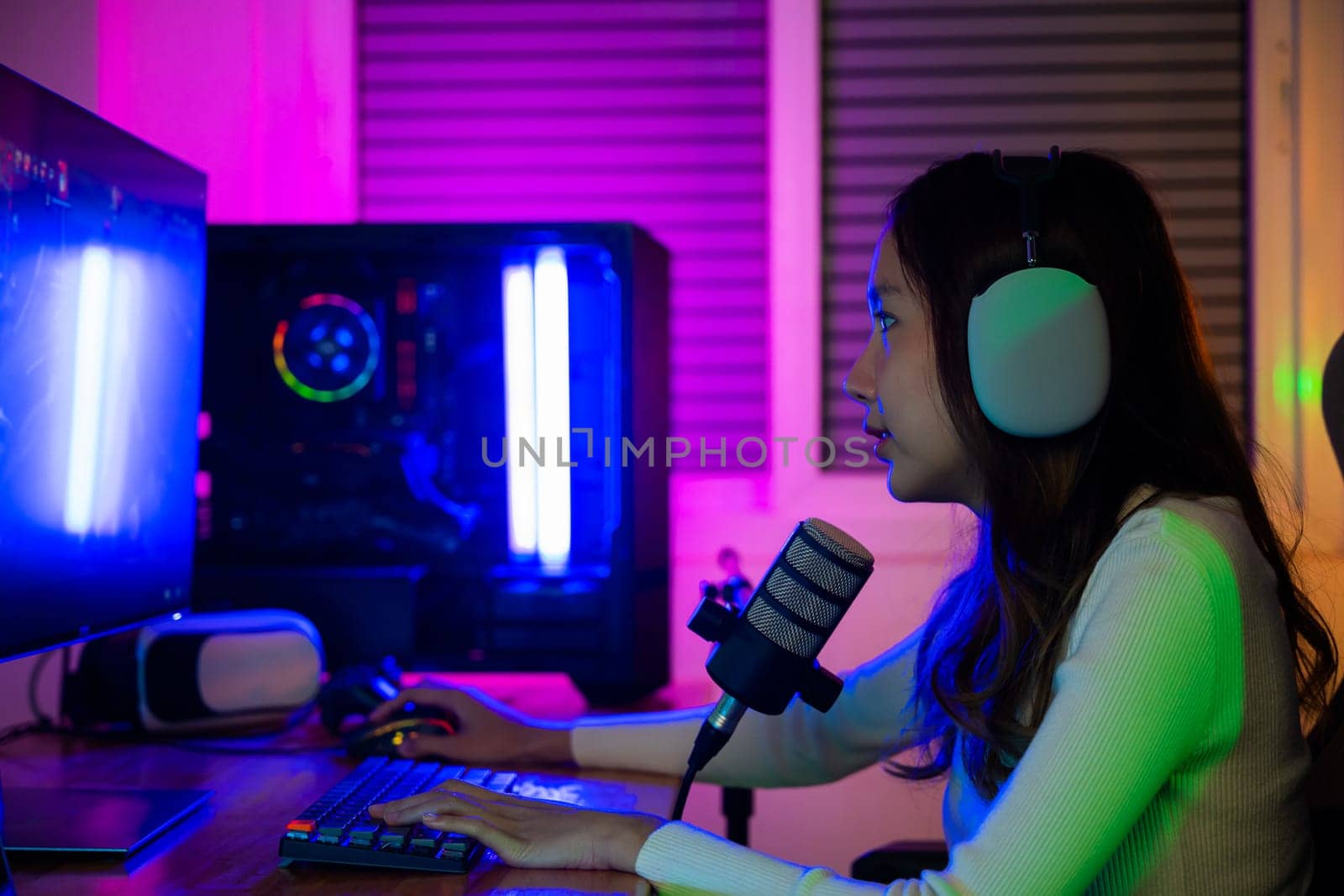 Asian young gamer woman wear headphones playing video games online on computer she live stream and chat with fans, Happy streamer female talking with microphone in gaming room, game streaming concept