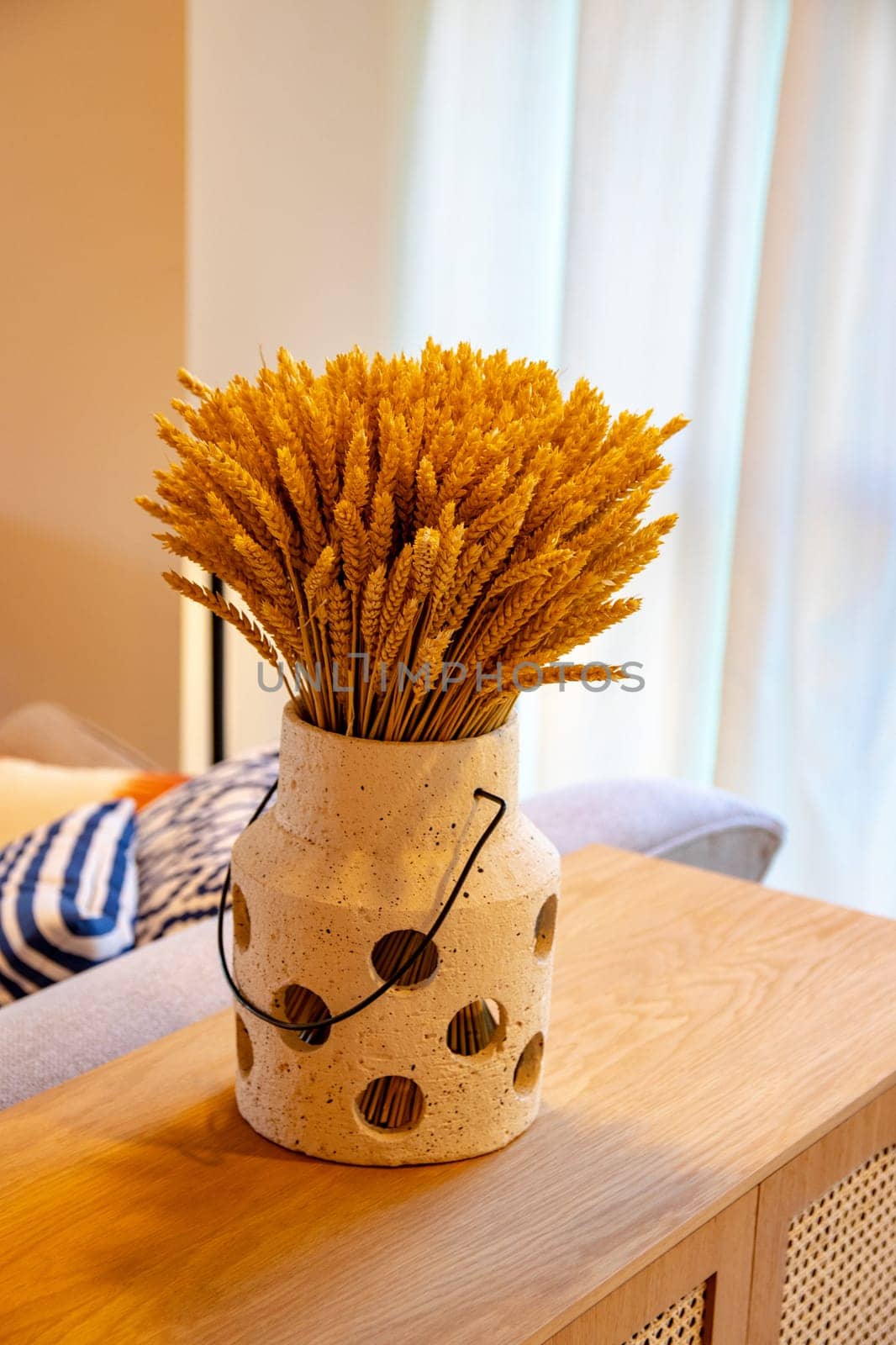 A sheaf or bouquet of dried wheat stalks stands in a vase on a wooden table against a light curtain background. Interior modern spring design. Dry flowers.