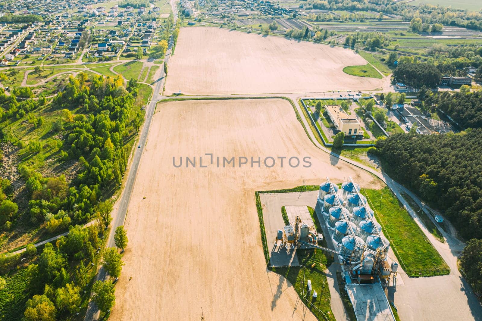Aerial View Modern Granary, Grain-drying Complex, Commercial Grain Or Seed Silos In Sunny Spring Rural Landscape. Corn Dryer Silos, Inland Grain Terminal, Grain Elevators Standing In A Field.