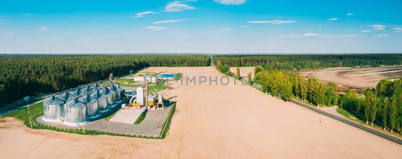 Aerial View Granary, Grain-drying Complex, Commercial Grain Or Seed Silos In Sunny Spring Rural Landscape. Corn Dryer Silos, Inland Grain Terminal, Grain Elevators Standing In A Field. Panorama.
