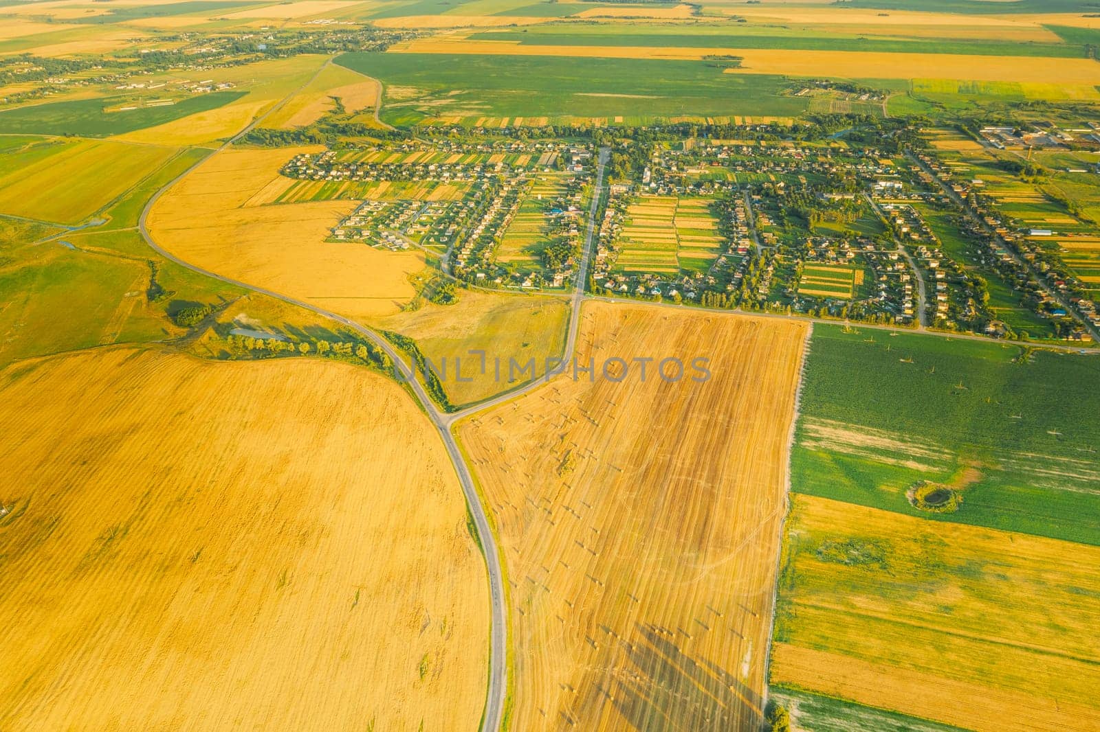 Aerial View Of Village And Hay Rolls, Straw Field Landscape In Summer Evening. Haystack, Hay Roll in Sunrise Time. Agricultural Countryside landscape In Harvest Season