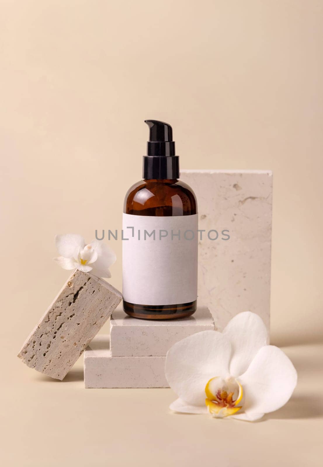 Dark glass one pump cosmetic bottle with blank label near white orchid flowers and stones on light yellow, close up, mockup. Skincare handmade beauty product, tonic or lotion. Exotic natural cosmetics..