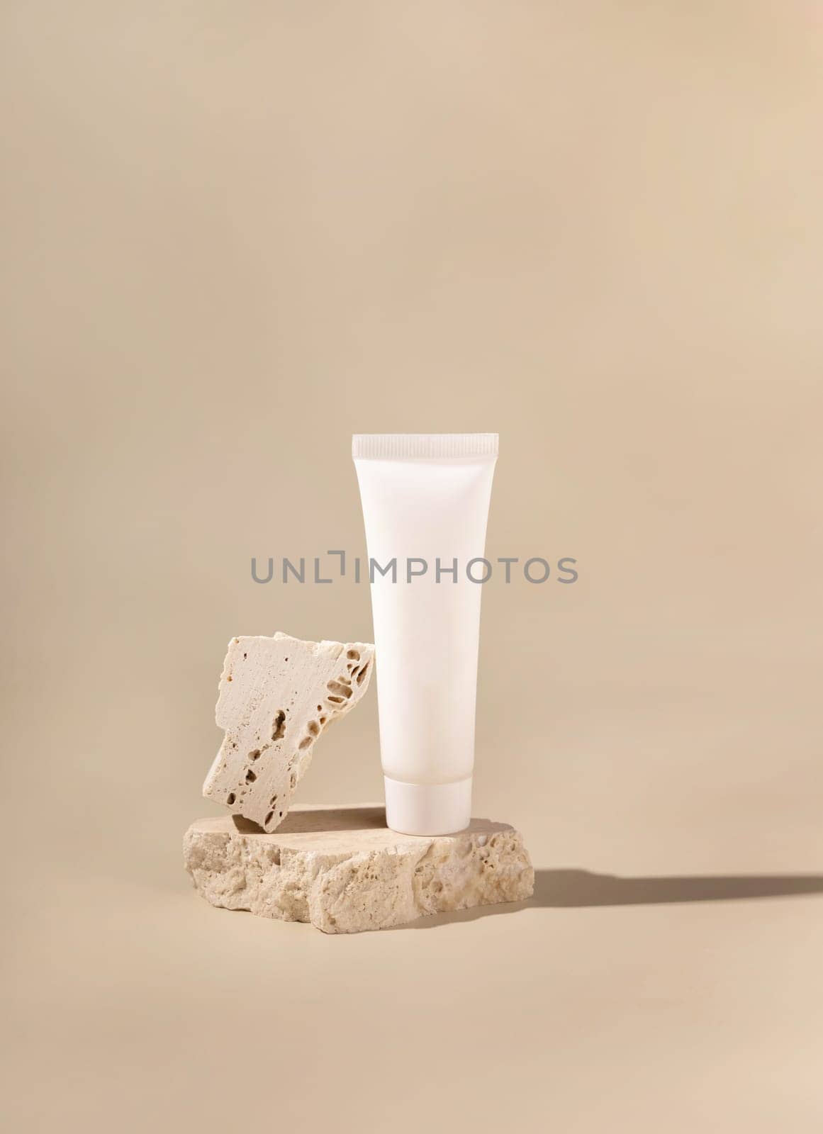 White plastic cream tube on travertine stone on light yellow, close up, mockup. Skincare beauty product. Natural cosmetics. Minimal composition..