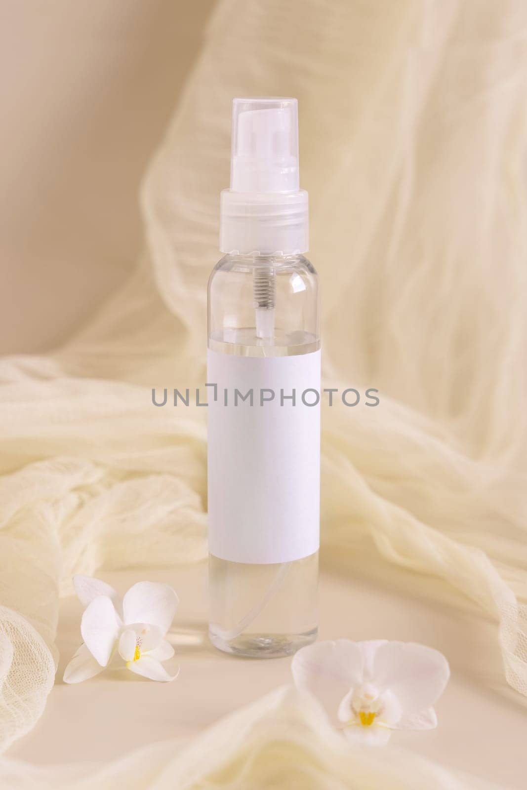 Spray bottle filled with liquid near orchid flowers and tulle on light beige close up. Packaging Mockup. Exotic natural cosmetics, romantic composition, everyday skincare routine..