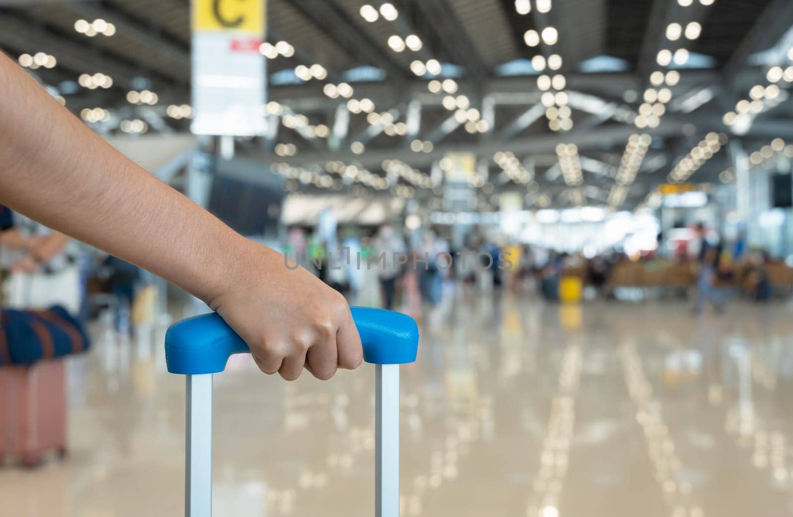 Woman traveler hand holding luggage with blurred passengers walking in airport terminal. Travel holiday and vacation concept.