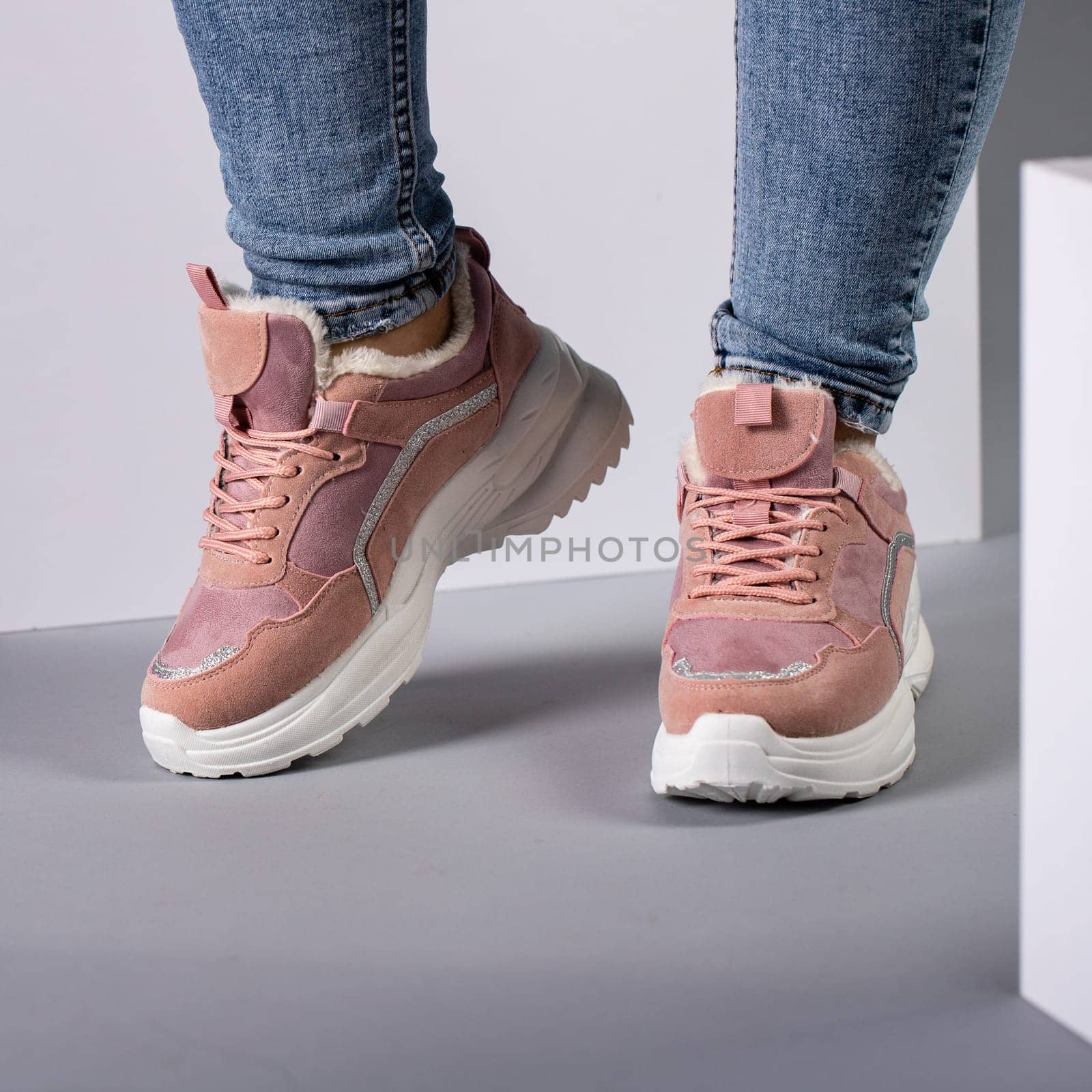 Cropped image of warm pink sneakers with down on female model legs in studio. Sports shoes on feet of unknown young woman who is dressed in jeans and stands on gray floor on background of white cubes.