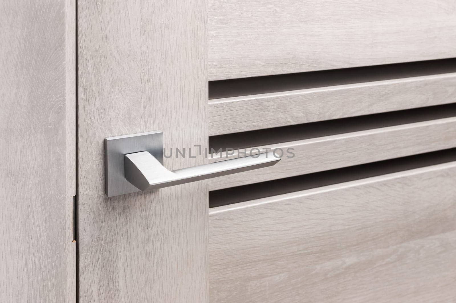 Close up of stylish metal door knob on modern interior door. Silver chrome door handle on light beige door with three strips of frosted glass. Concept of interior details.