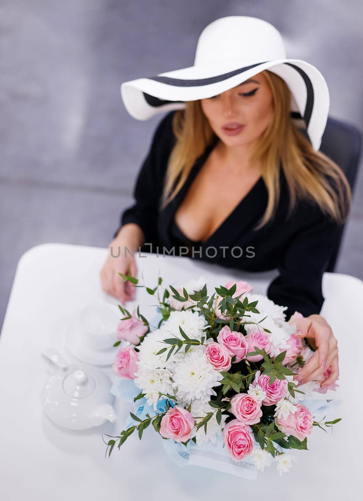 Bouquet of white chrysanthemums and pink roses on table in restaurant near glamorous woman. Top view of woman in white hat elegantly touches flowers sitting at table and drinking tea. Selective focus.