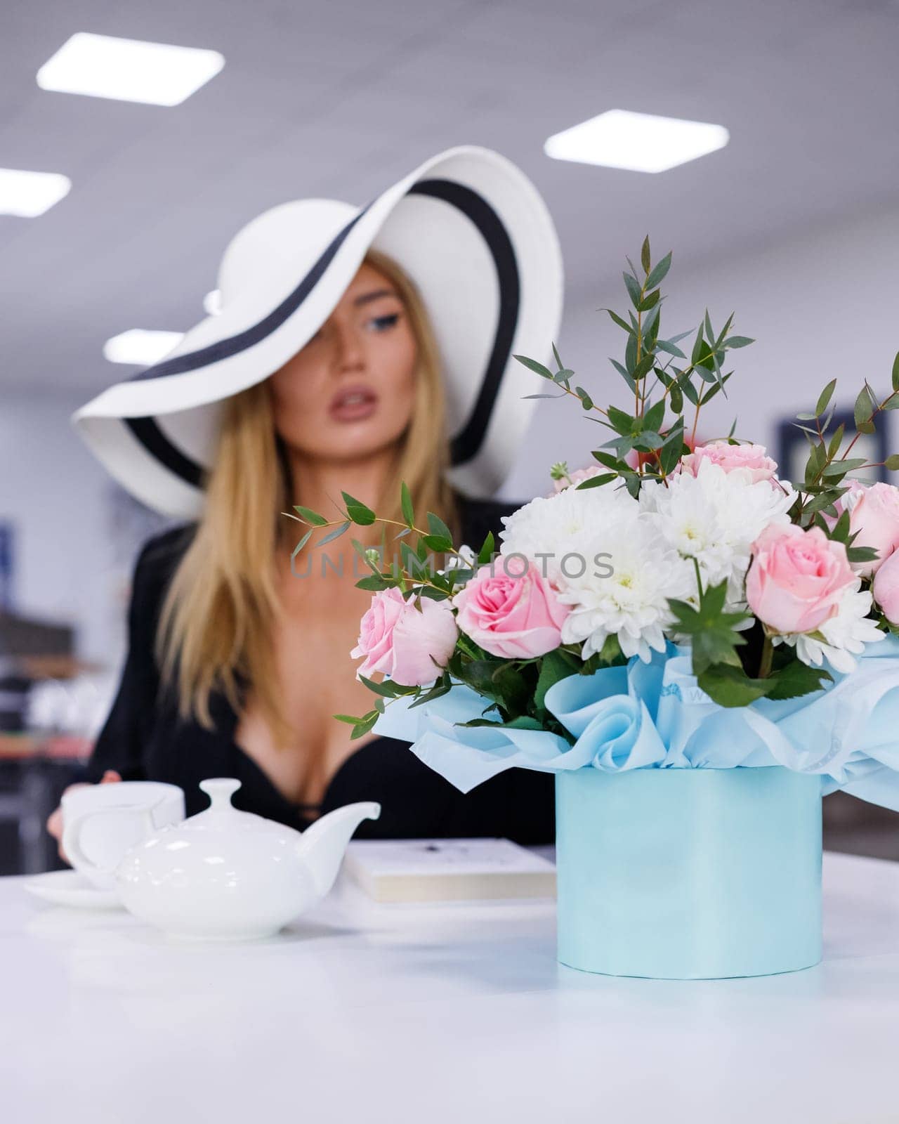 Fashion model portrait. Stylish beautiful young woman resting in restaurant sitting near bouquet of flowers. Woman with make-up dressed in white hat sits at table with teapot, cup and box with flowers.