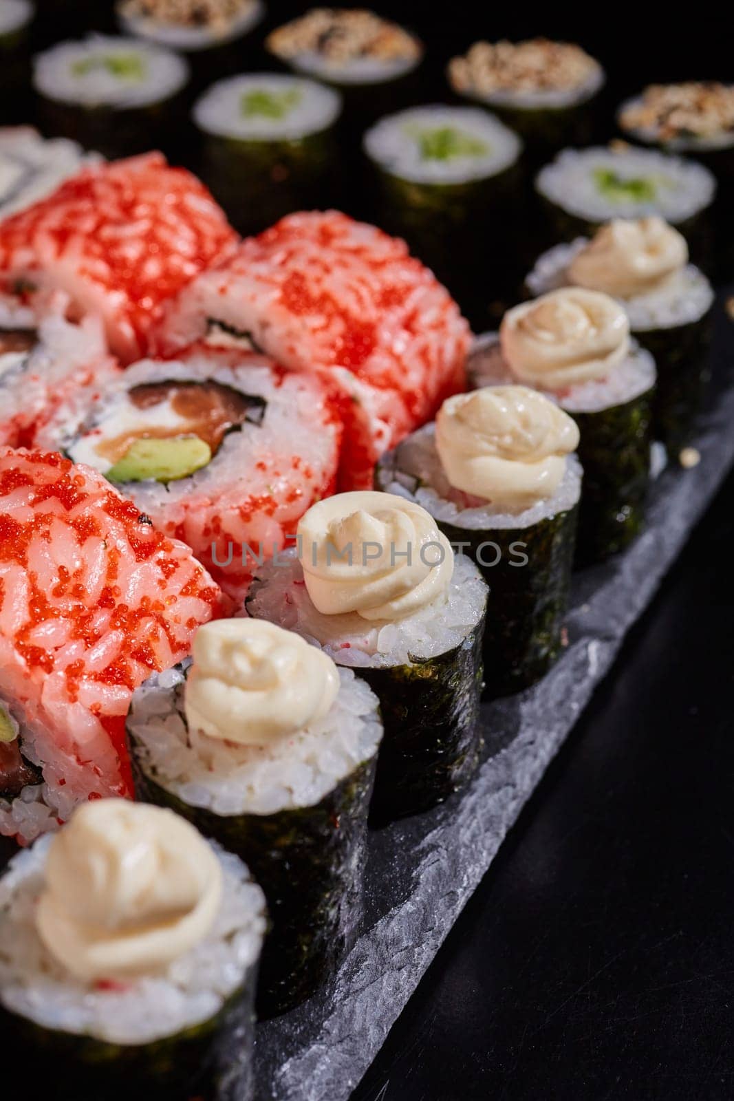 Close up of maki rolls with cream cheese lying on a black flat plate around california rolls with caviar. Japanese food concept.