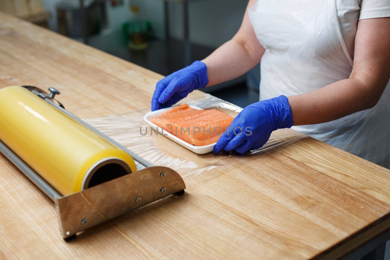 Worker in factory or shop packs raw fresh salmon fillets in cling film. Unknown worker in blue disposable gloves packs fish lying in white tray unwinding film from roll in front of it.
