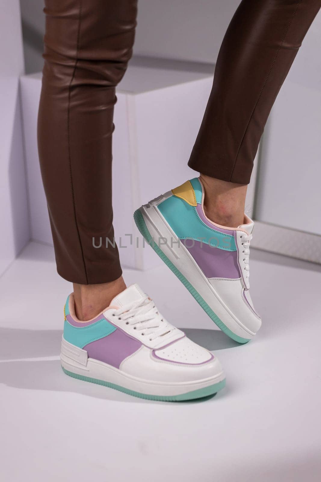 Close up of stylish modern female sneakers on model legs in studio. Side view of white sneakers with turquoise and purple inserts dressed on woman in brown pants standing on light background.