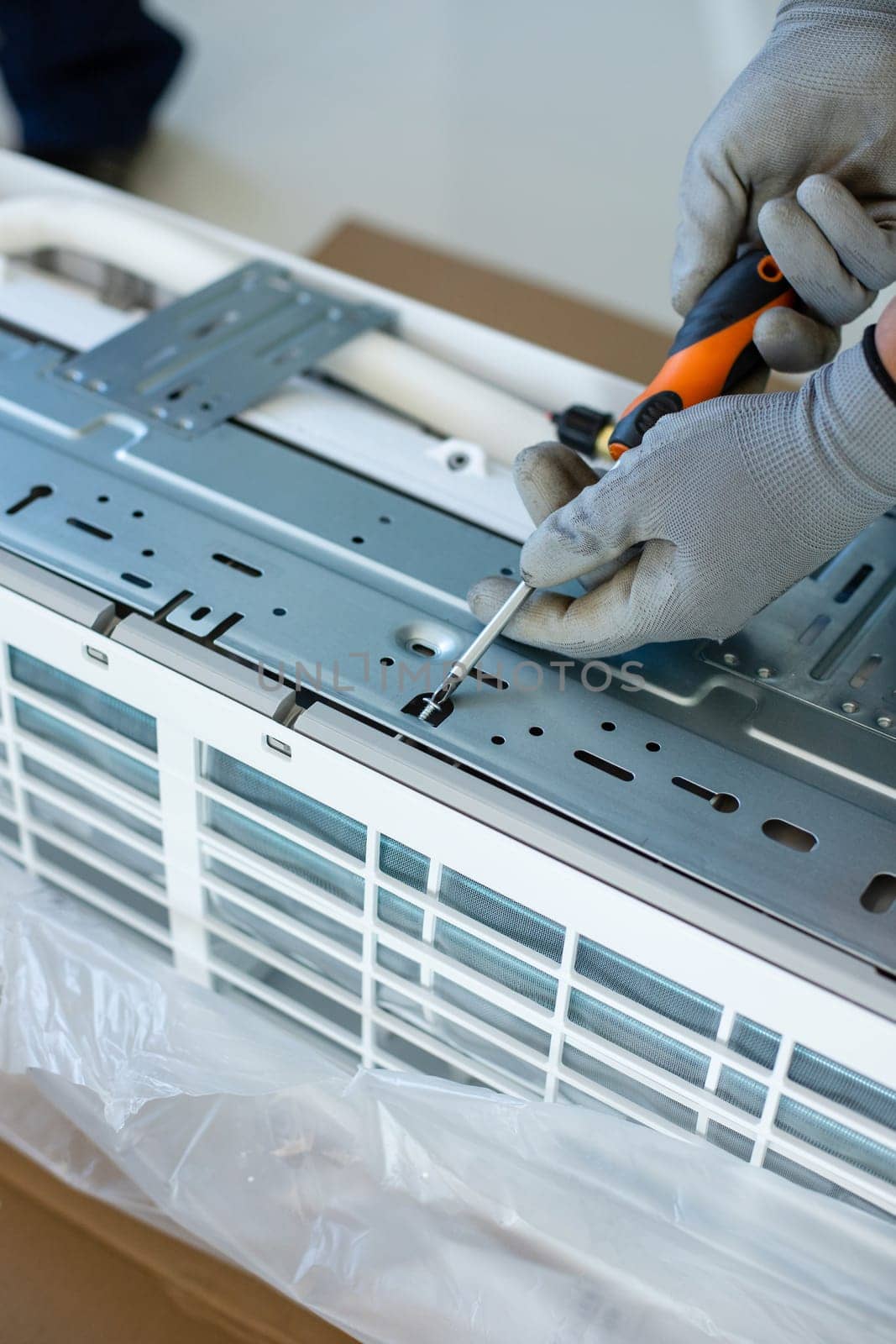 Male installer or repairman is going to install air conditioner unit. Close up of hands of young skilled male worker in household gloves who fastens bolts on air conditioner with screwdriver.