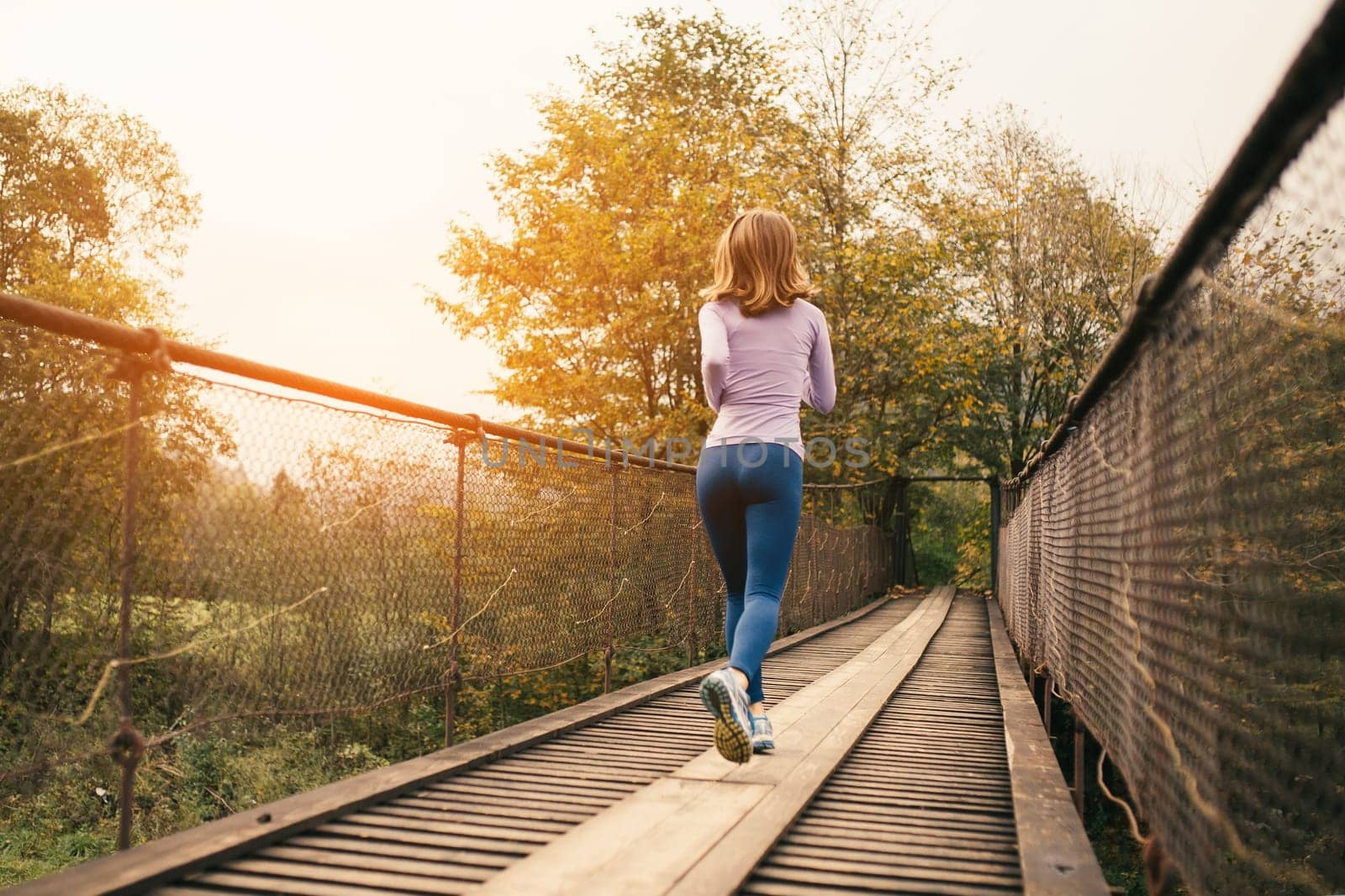 Fitness and sports motivation. Rear view of a sporty woman runner running over an old suspension bridge on a cool autumn day. Outdoor cardio exercise concept.