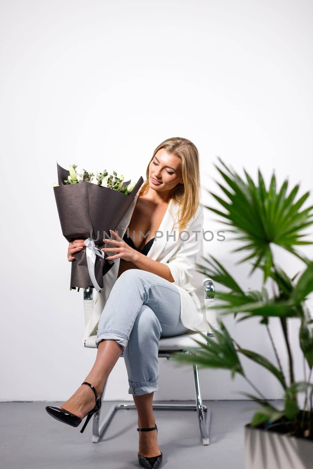 Spectacular young blonde woman with bouquet of flowers in her hands. Woman sitting on chair near green flowerpot on white studio background and looking at bouquet that is wrapped in paper.