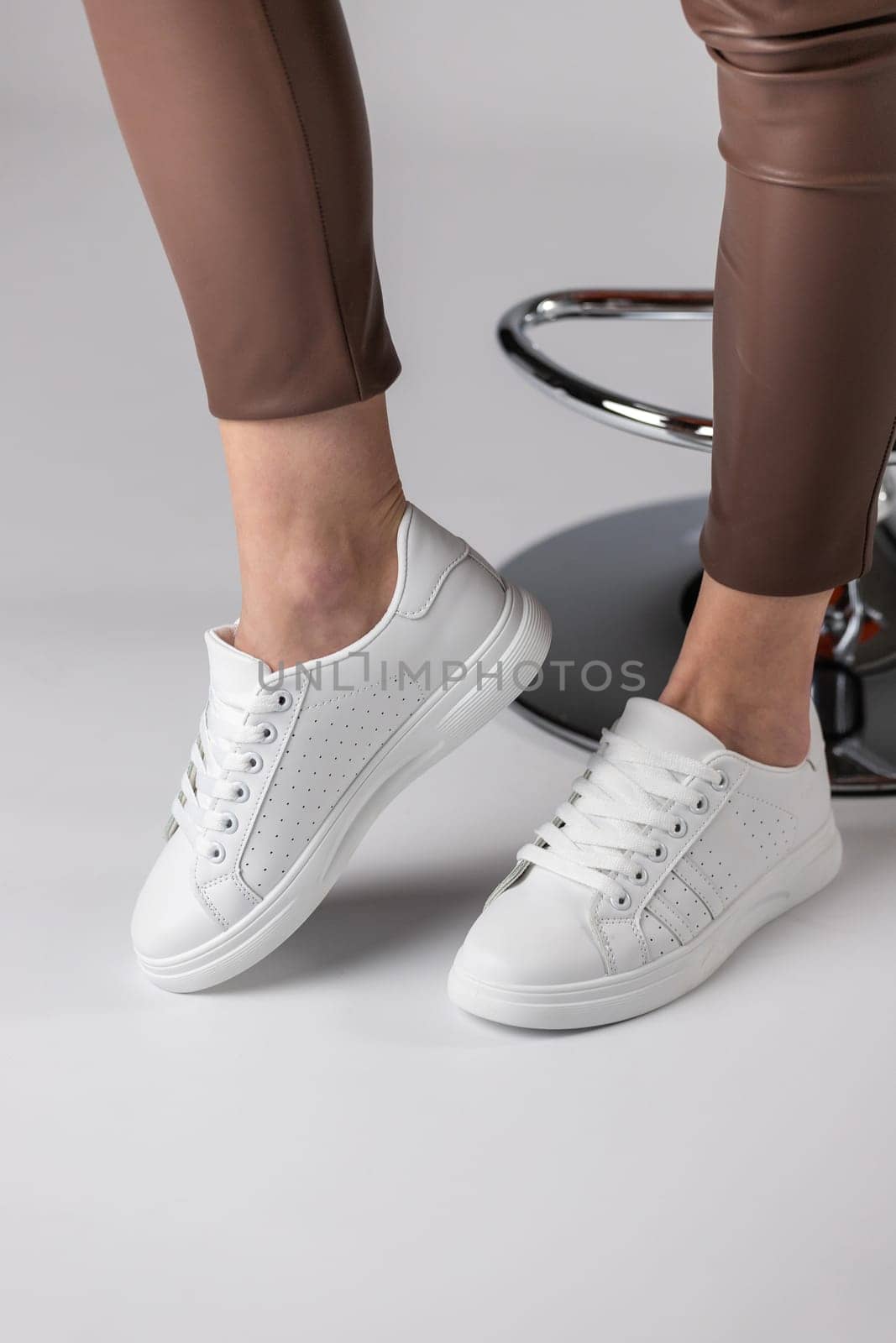 Close up of fashionable white sneakers on legs of girl on white background in studio. Young woman in brown pants demonstrates new collection of sports shoes with laces and with breathable holes.