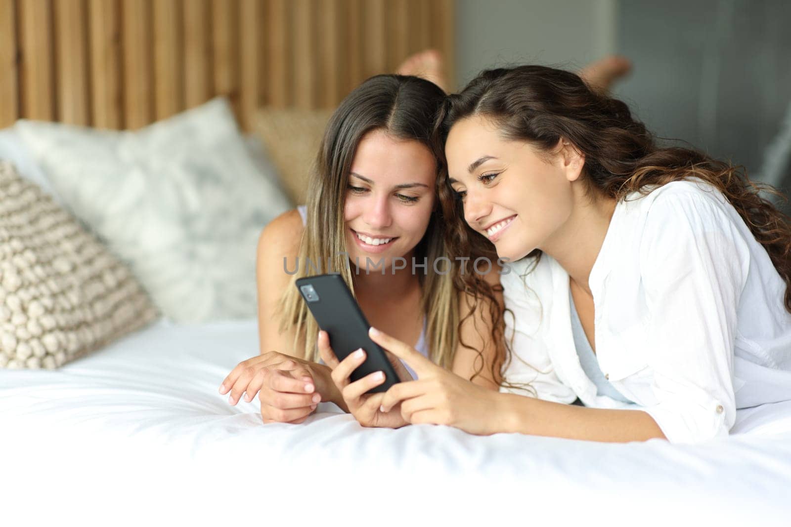 Two happy friends checking smartphone lying on a bed