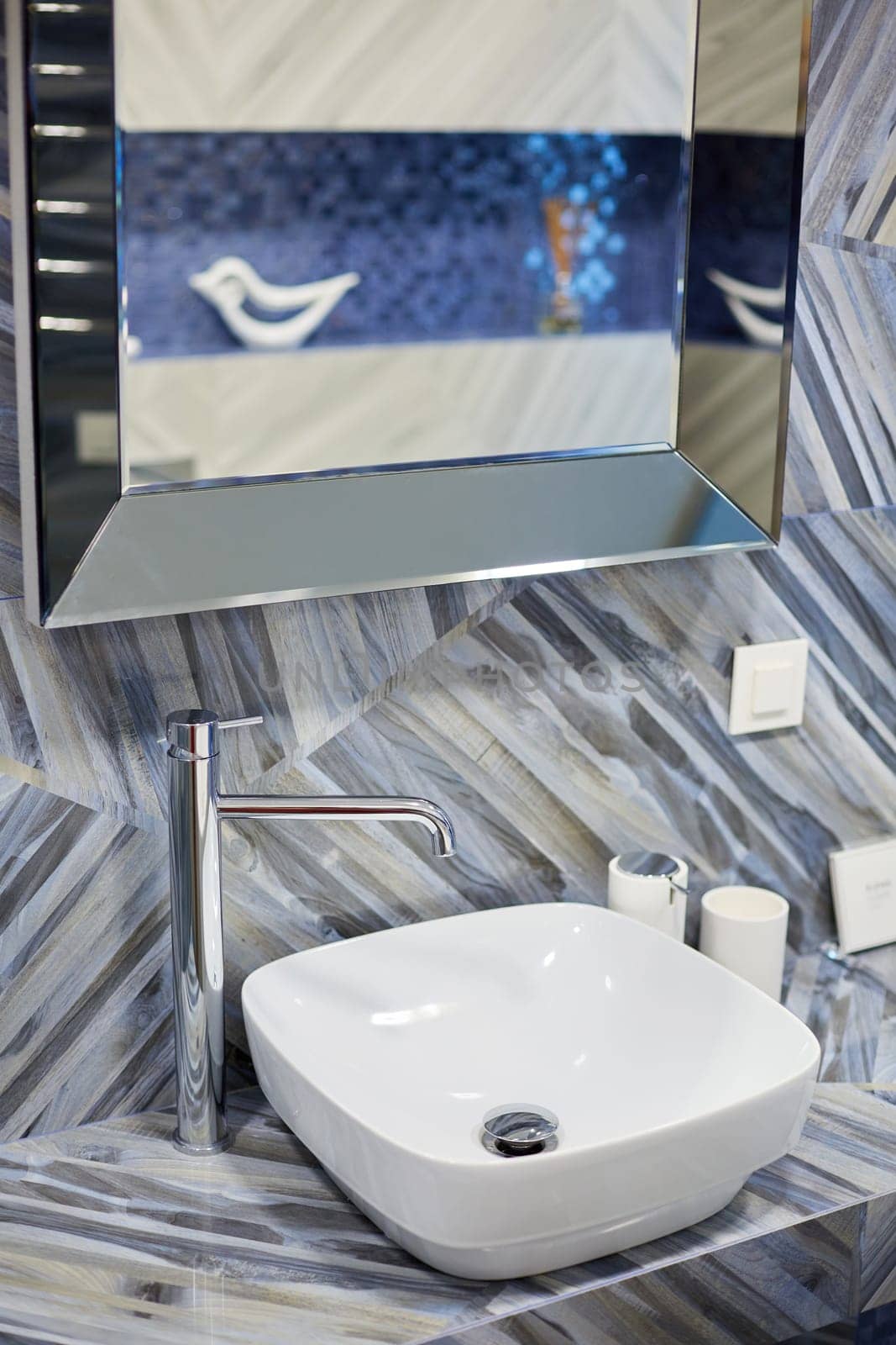 Modern washbasin with chrome faucet in stylish and bright bathroom of new house. Mirror hangs over sink in room with trendy tiles on walls. Concept of details.