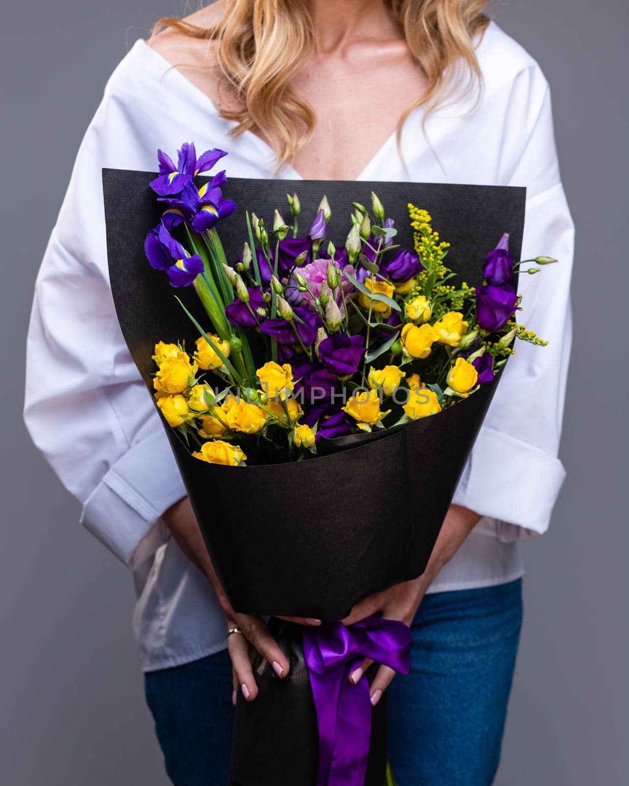 Stylish bouquet of yellow roses and purple eustoma in the hands of a blonde woman standing on a gray background. Concept of congratulations on International Women's Day, Birthday and Mother's Day.