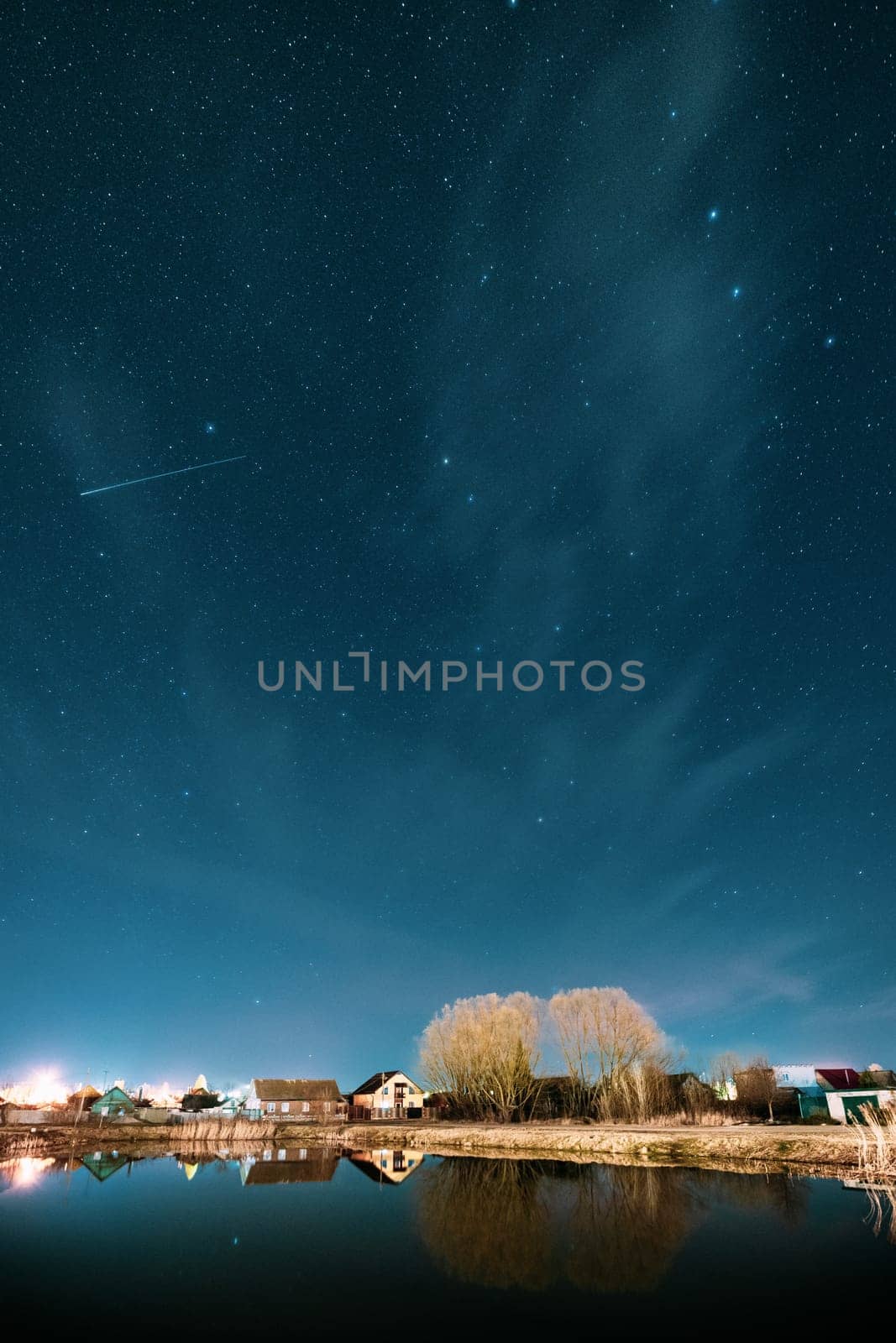 Belarus, Eastern Europe. Night Sky Stars Above Countryside Landscape With Lake Coast And Small Town Or Village. Natural Starry Sky Above Pond And Houses In Early Spring Night.