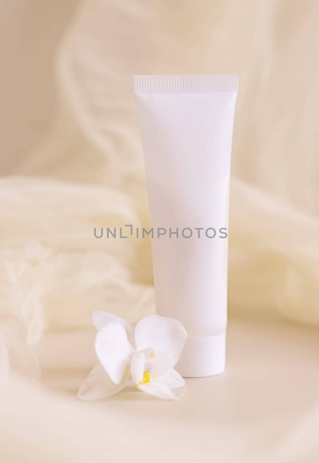 White Cream tube near orchid flowers and tulle on light beige close up. Packaging Mockup. Exotic natural cosmetics, romantic composition, everyday skincare routine..