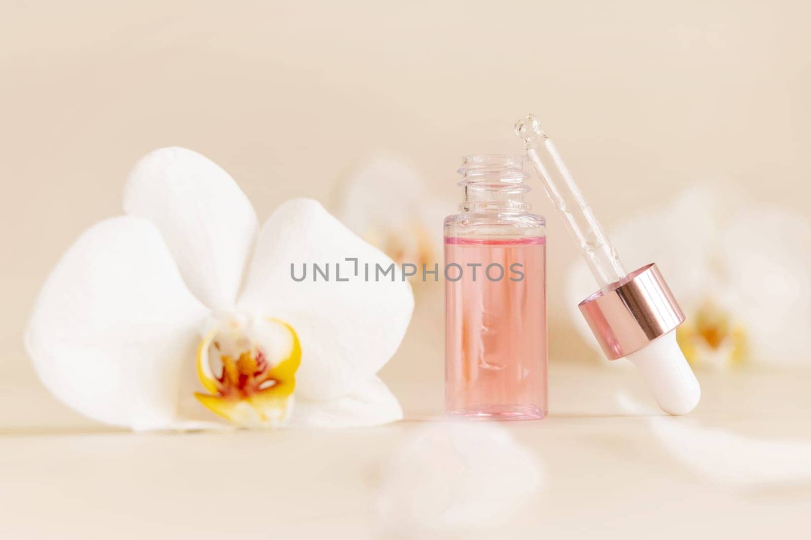 Light Pink opened Dropper Bottle with serum and a pipette cap near white orchid flowers on light beige, close up, copy space. Skincare cosmetic product, essence or serum. Everyday beauty routine.