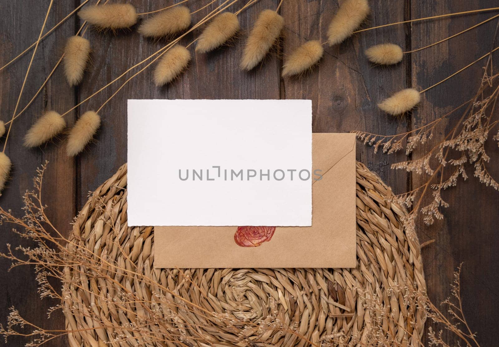 Blank card and envelope on wattled placemat on wood near Hare's Tail grass top view. Boho scene with horizontal paper card. Bohemian or close to nature greeting or Invitation mockup, place for text.