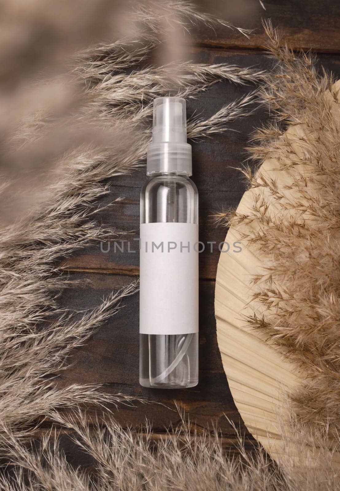 Transparent spray bottle on wood near dried palm leaf and pampas grass top view, label mockup. Skincare beauty product, cleanser or water. Bohemian eco friendly cosmetics flat lay with dried leaves.