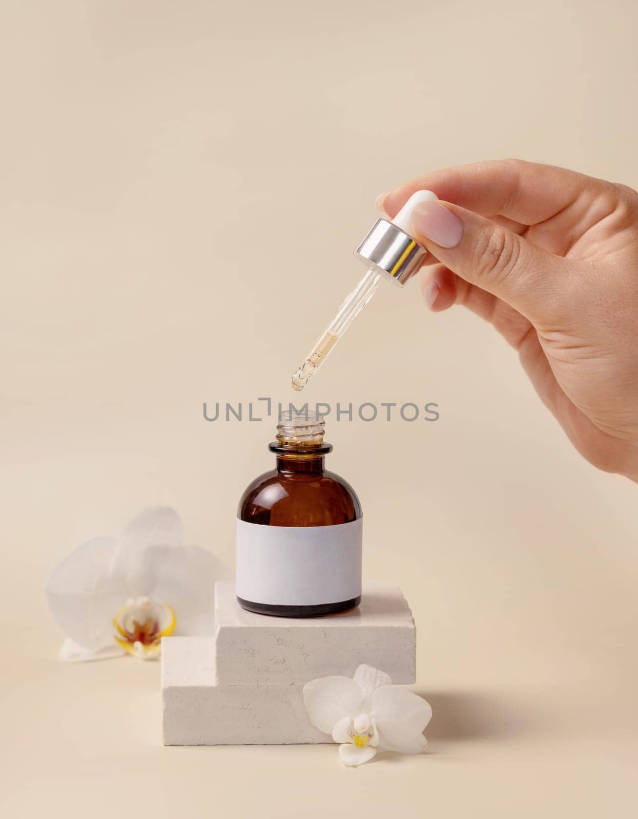 Hand holding pipette over opened cosmetic dropper bottle near white orchid flowers and stones on light beige, close up, mockup. Natural skincare product, serum or essential oil.