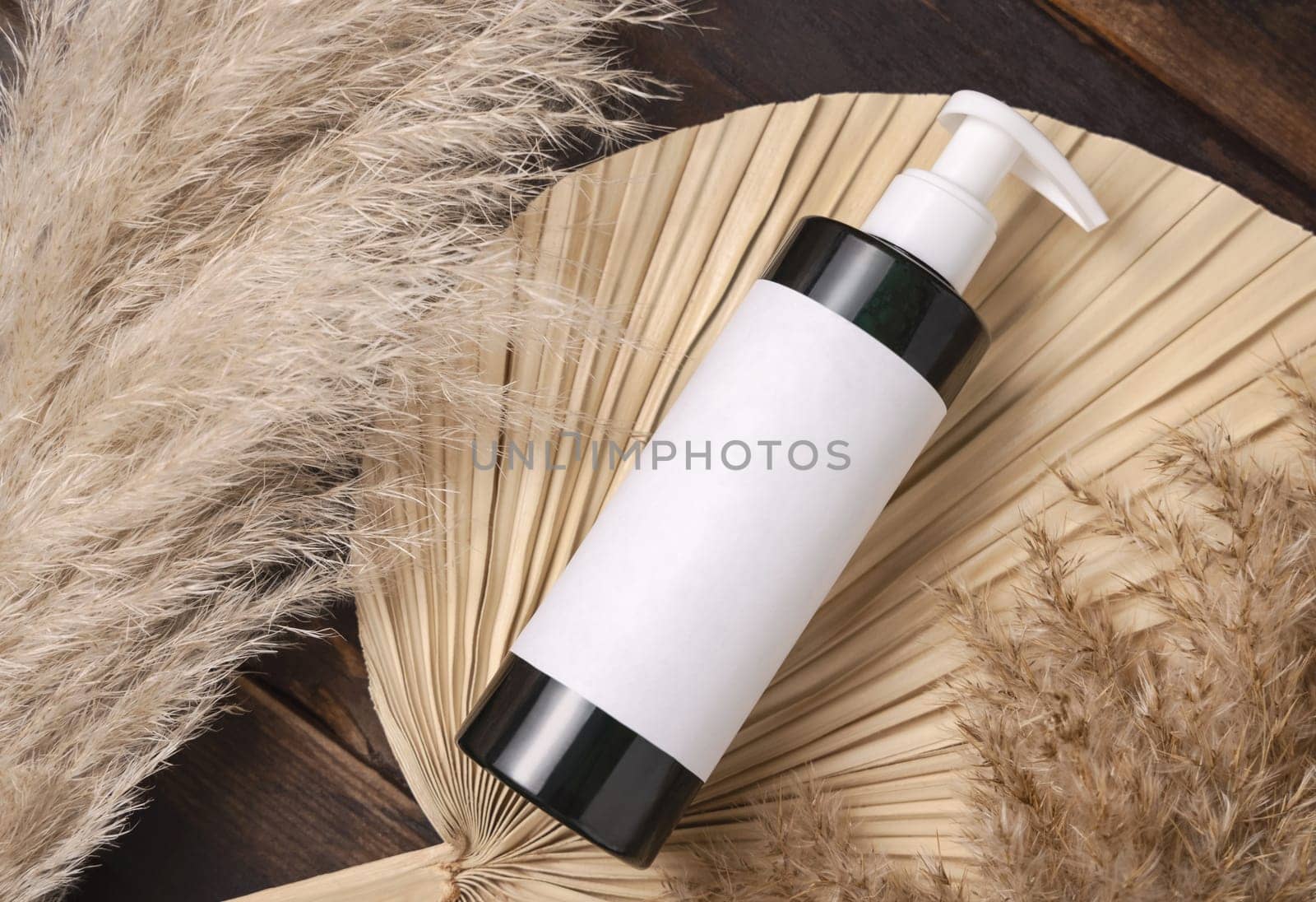 Cosmetic one pump bottle on dried palm leaf near pampas grass top view, label mockup. Skincare beauty product, lotion or essence. Bohemian eco friendly cosmetics flat lay with dried leaves.
