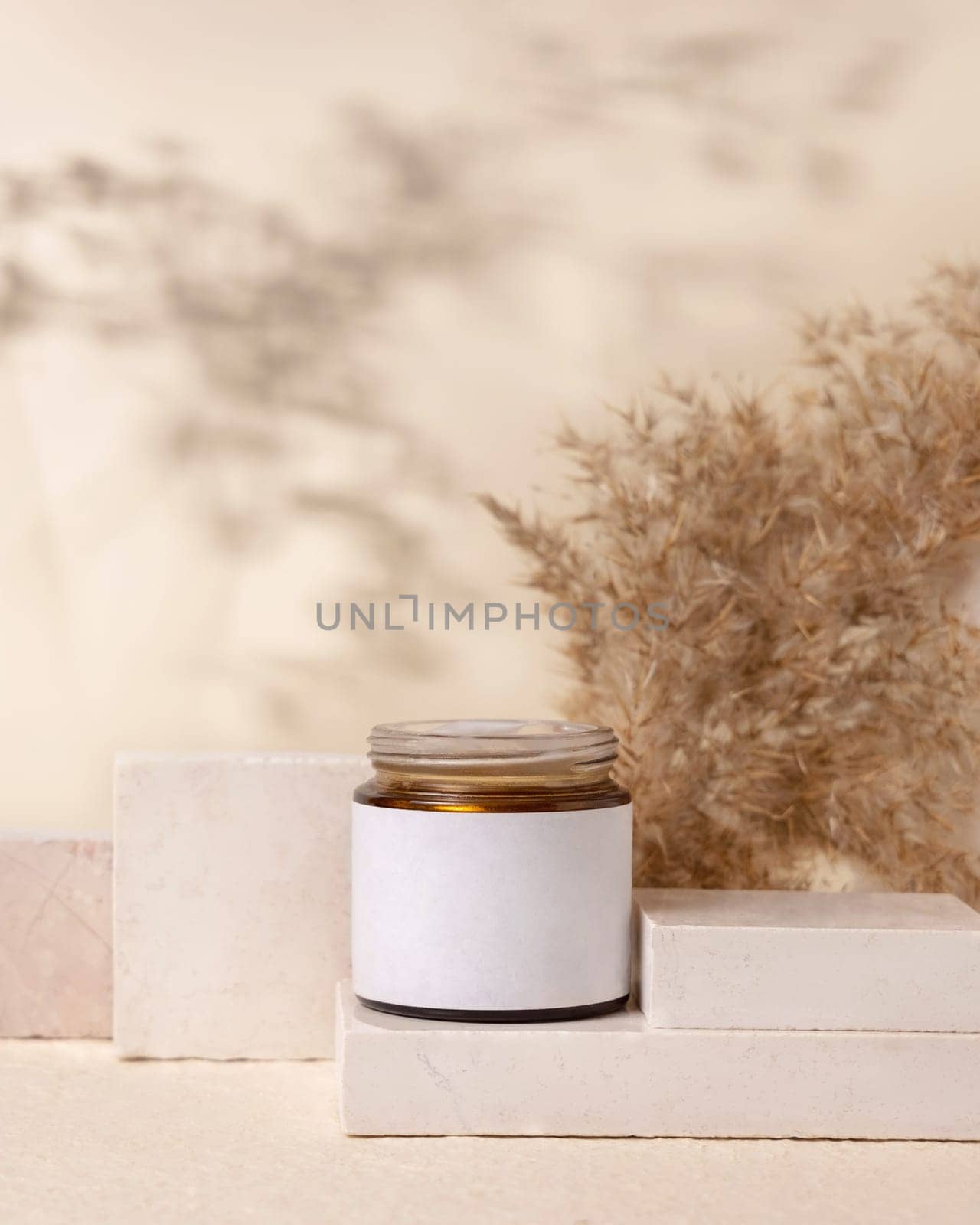 Glass cosmetic jar with blank label on stones near pampas grass on light beige close up, packaging mockup. Skincare beauty product, lotion or cleanser. Natural eco friendly cosmetics