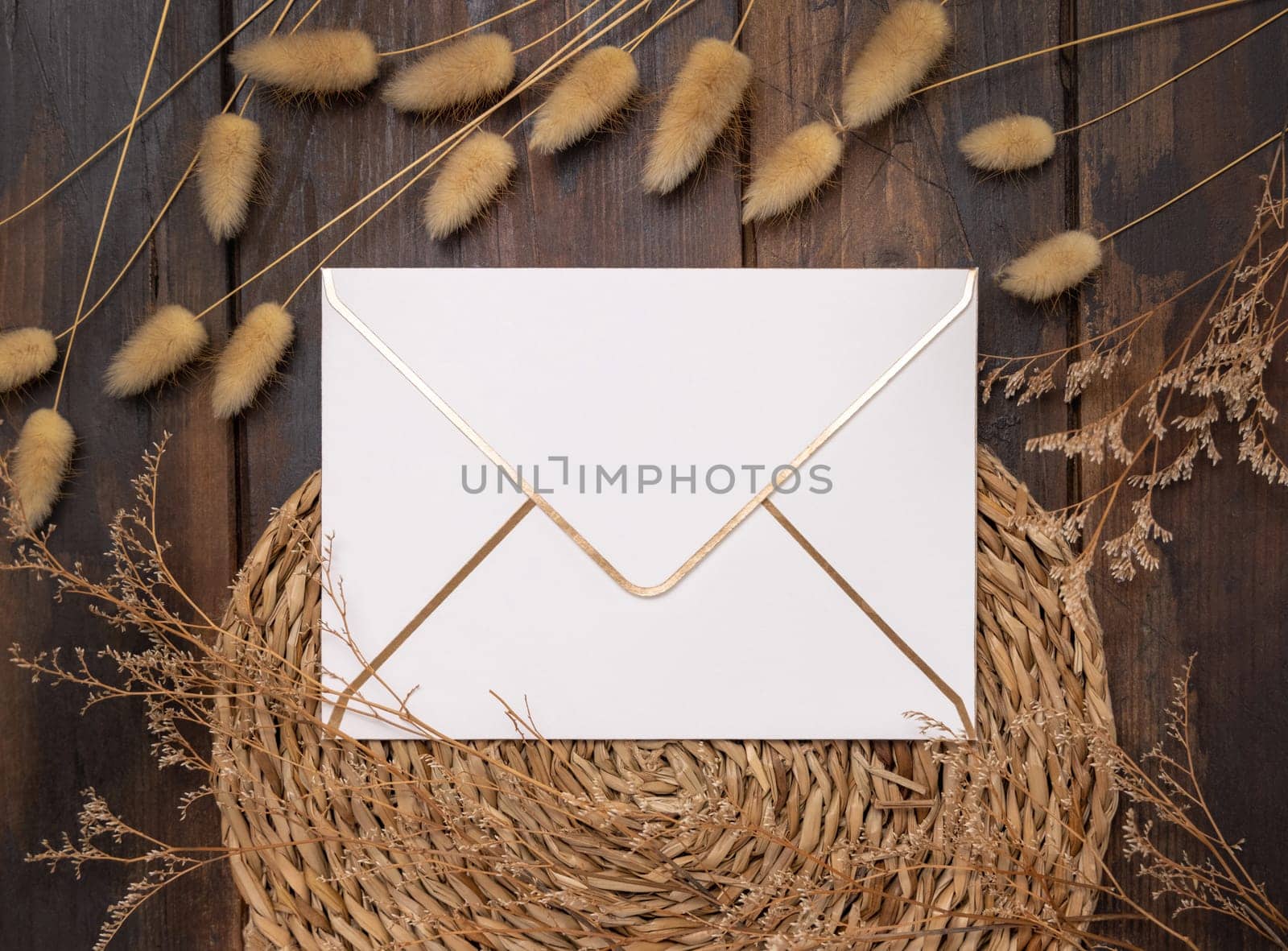 Blank envelope on wattled placemat on wood near Hare's Tail grass top view. Boho scene with horizontal paper envelope. Bohemian or close to nature greeting or Invitation mockup, place for text.