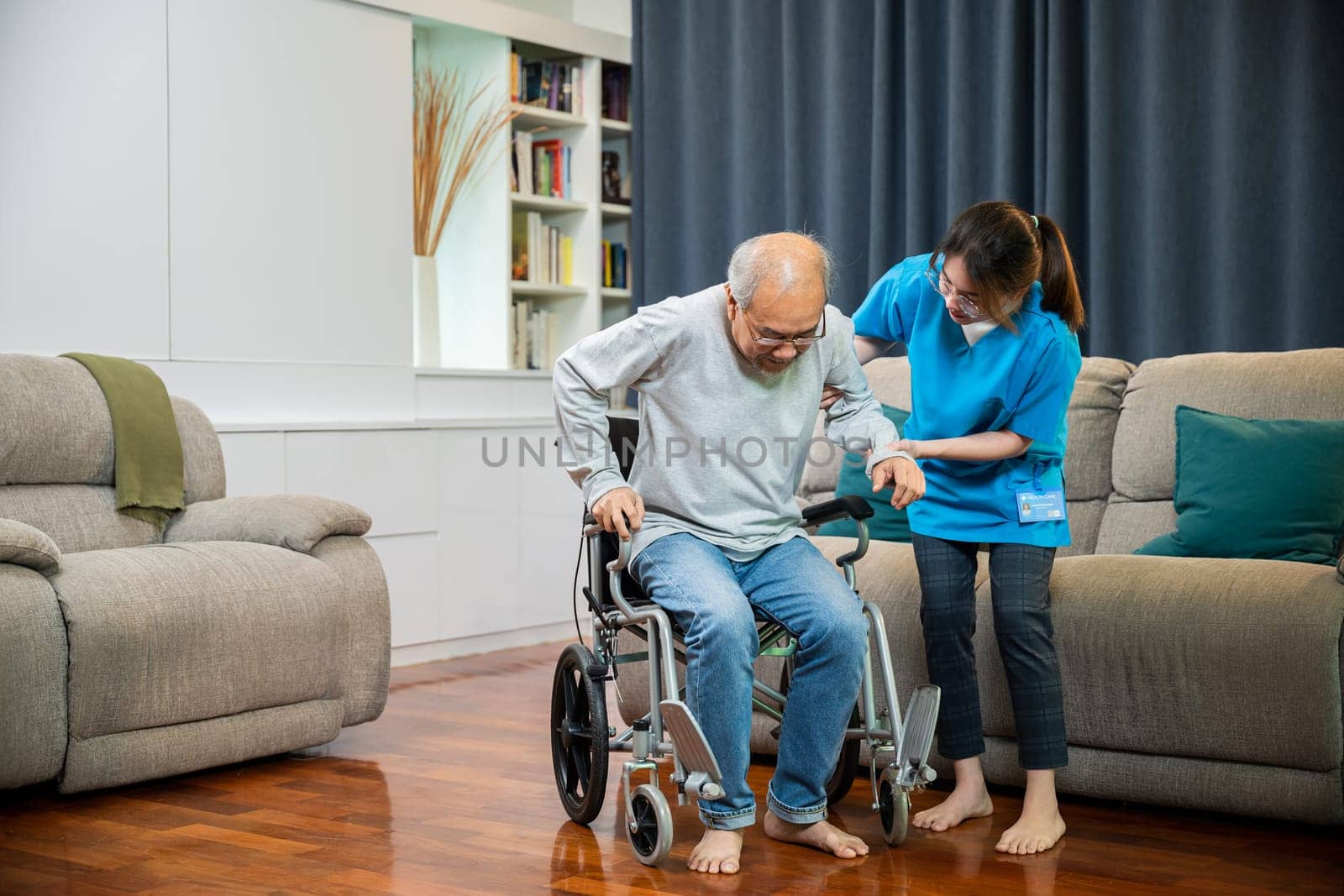 Asian nurse assisting helping senior man patient get up from wheelchair for practice walking at home, Smiling doctor support old man to getting up, help handicapped elderly stand up, healthcare
