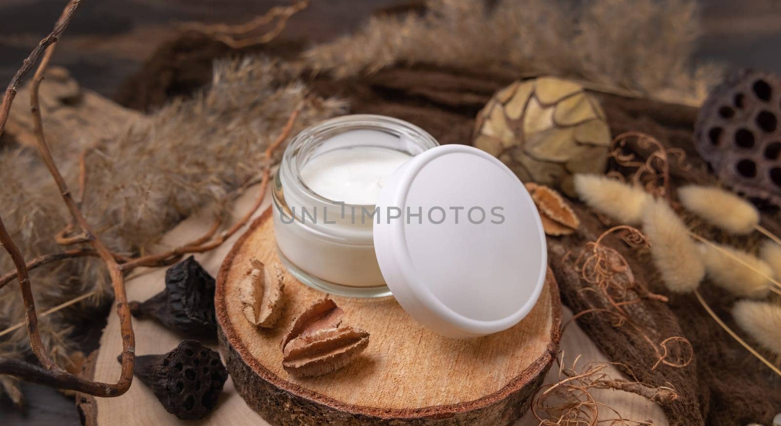 Opened glass cream jar with blank lid on wood near natural decorations close up, package mockup. Eco friendly skincare product, lotion or cream. Bohemian composition with dried leaves and flowers.