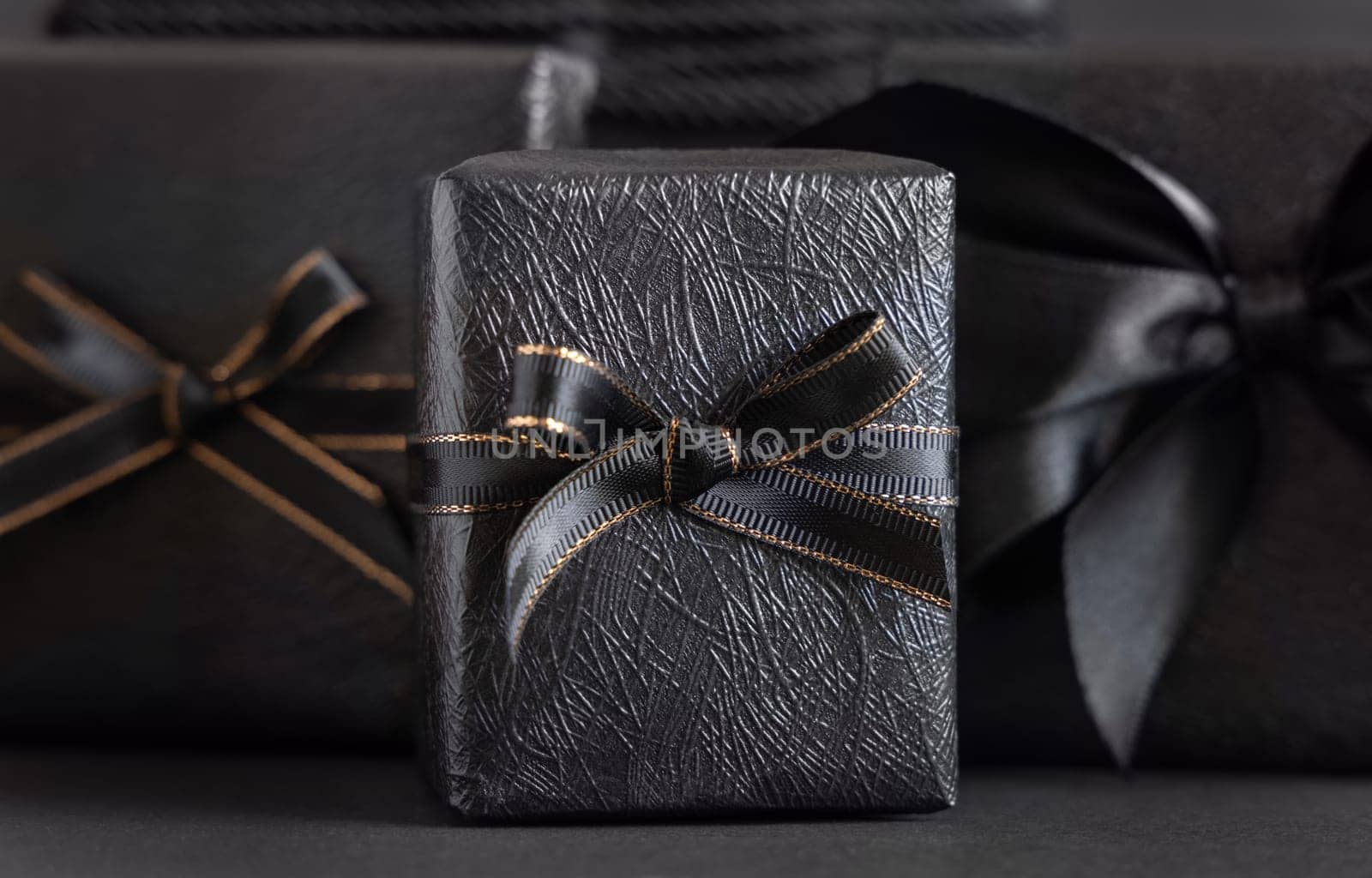 Black Gift Boxes with a ribbon bow on black close up. Wrapped presents for Dark themed Party, Christmas, Birthday, Anniversary. Black Friday concept. Monochrome composition