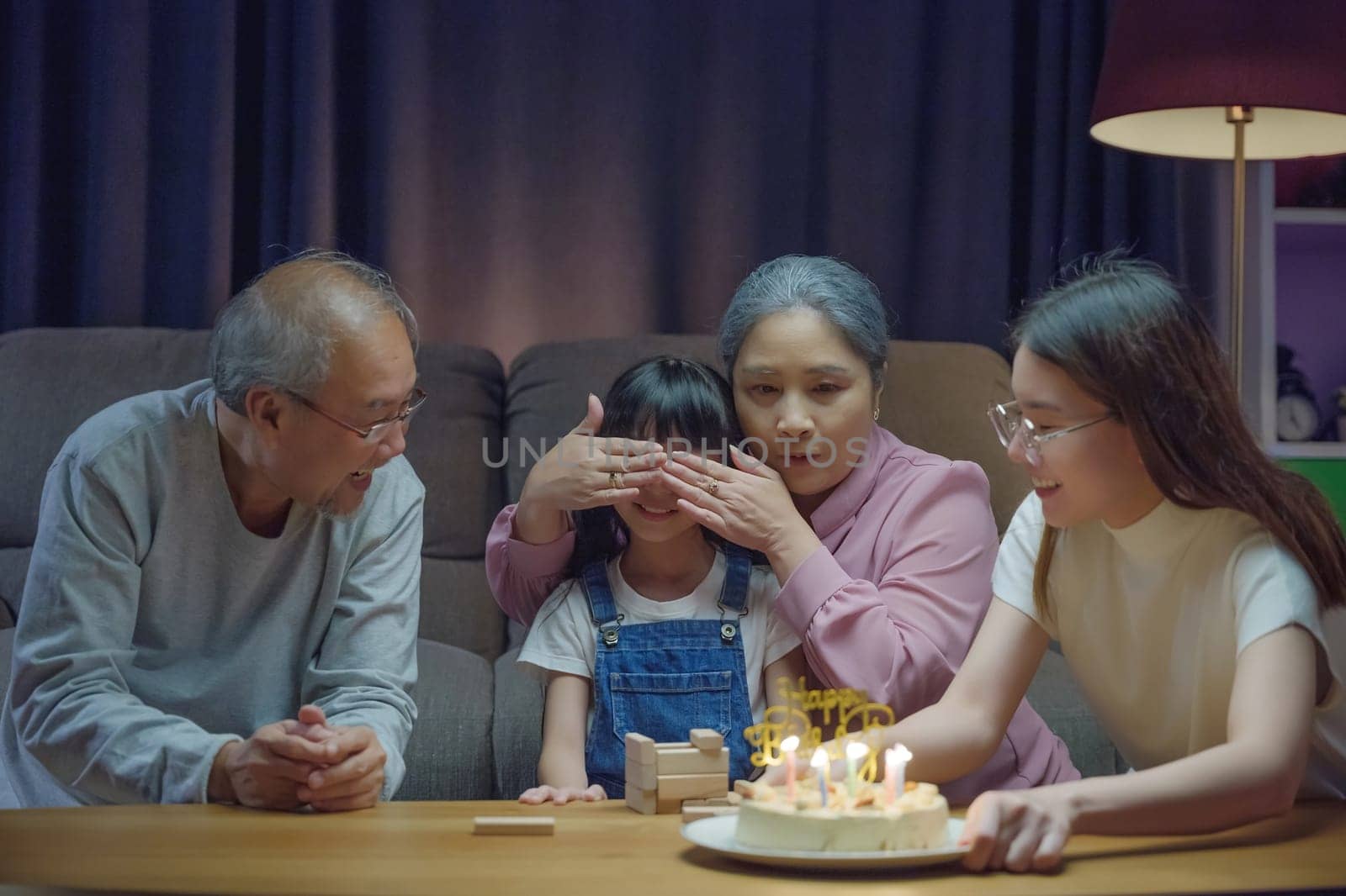 Birthday surprise concept. Happy Asian family grandparents and mother celebrating enjoy singing birthday song party with cake of granddaughter blowing candle at home in night time