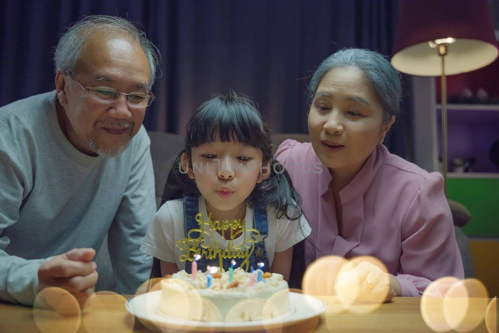 Happy Asian family grandparents and mother celebrating enjoy birthday party with cake of granddaughter Blowing candle at home in night time, Birthday surprise concept