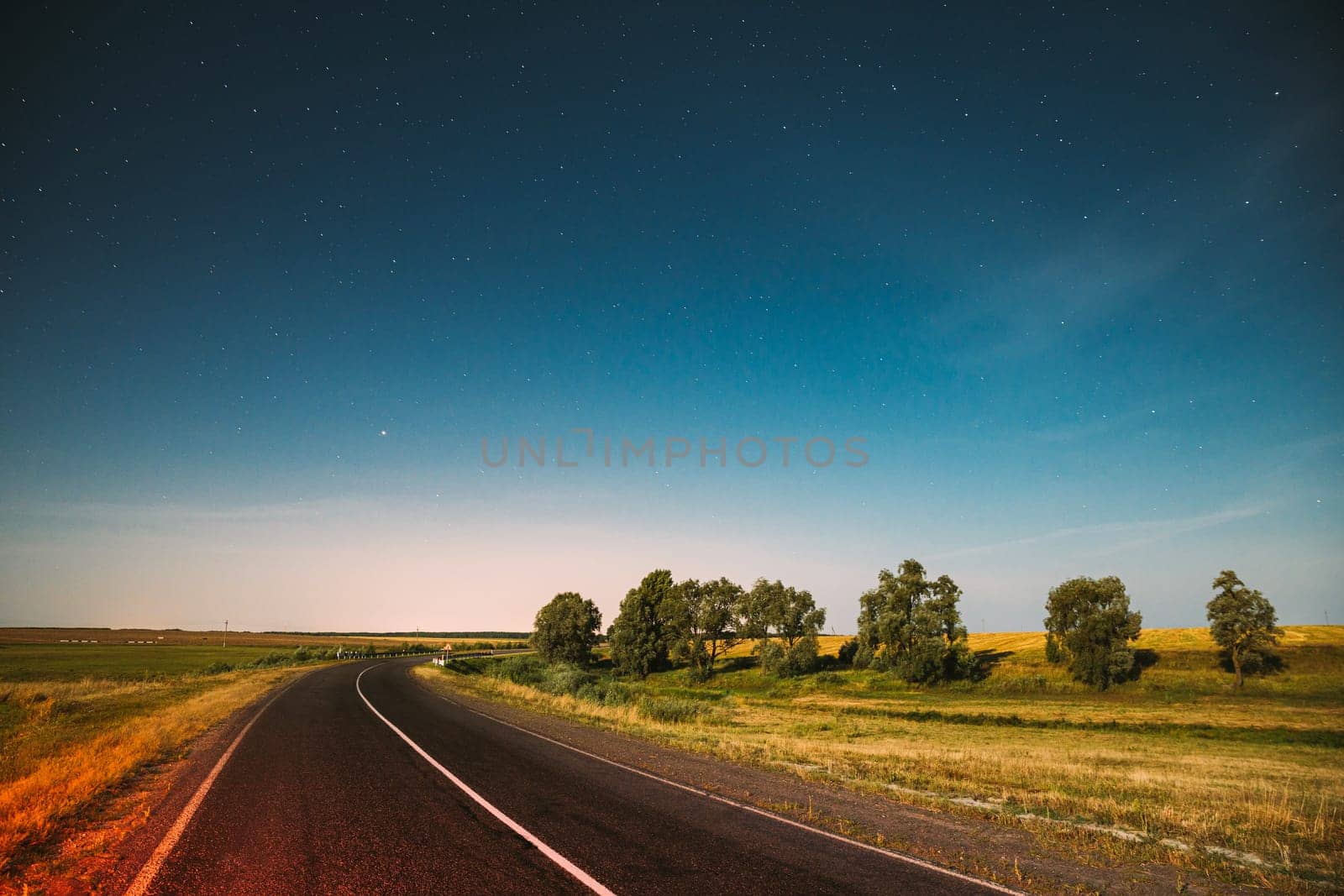 Blue Night Starry Sky Above Country Asphalt Road In Countryside And Green Meadow. Night View Of Natural Glowing Stars.